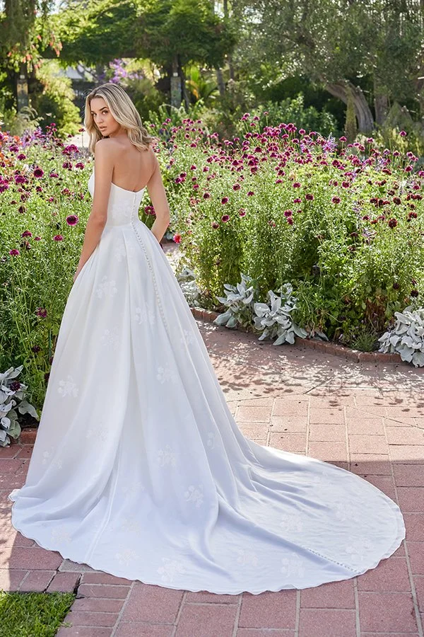 Rear view of a bride in a modern strapless A-line wedding dress featuring a clean straight back, subtle floral jacquard fabric, covered buttons flowing into a graceful chapel train, photographed in a beautiful flower-filled garden.