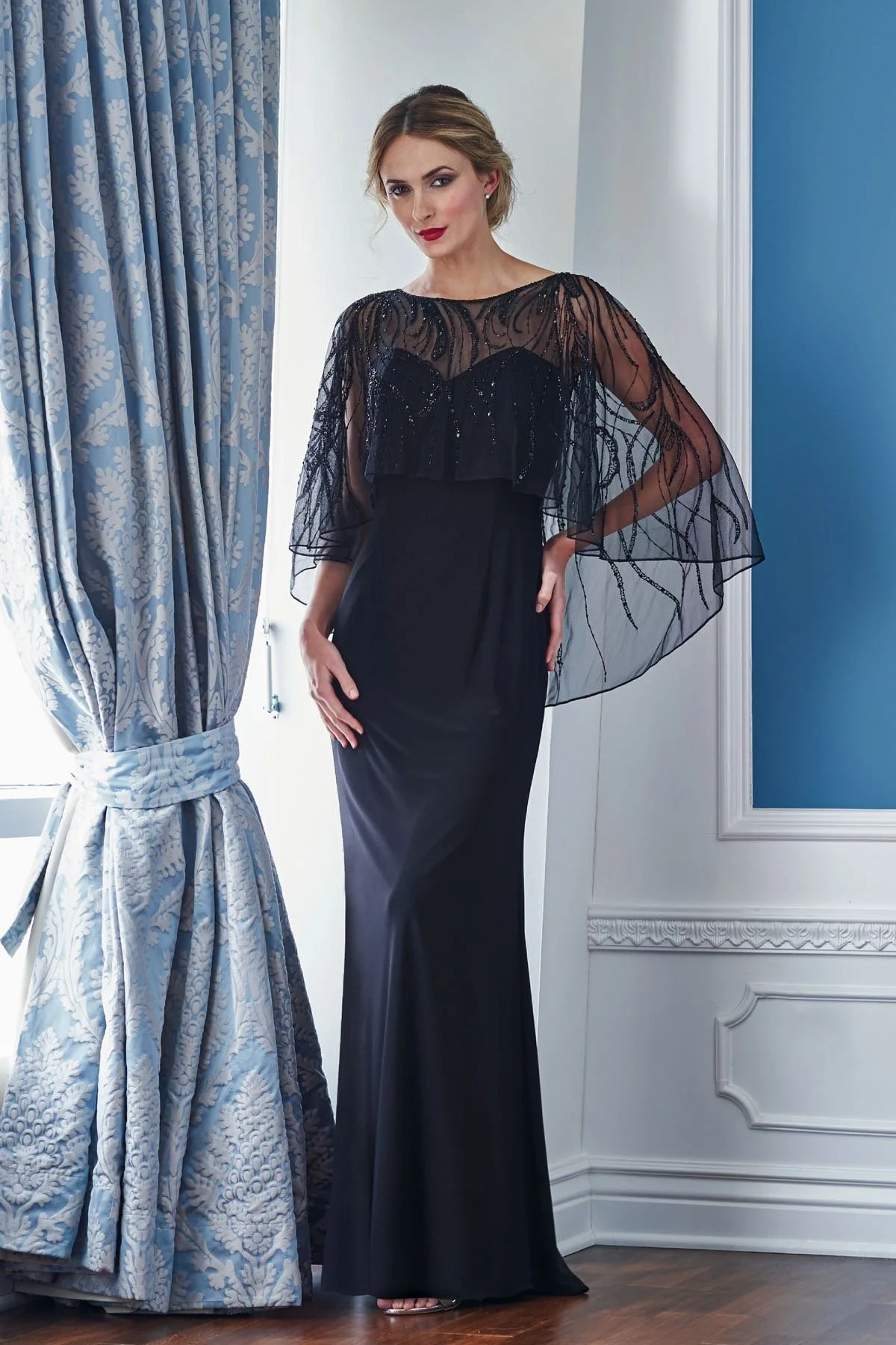 Front view of a black mother of the bride gown with an illusion beaded bodice overlay, dramatic cape sleeves, and a sleek floor-length silhouette for elegant weddings and formal events.