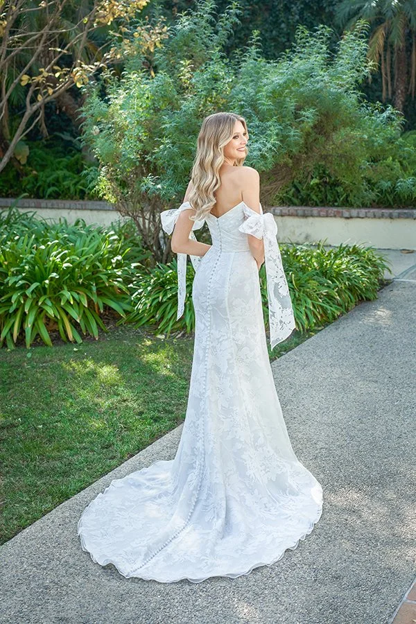 Back view of an off-the-shoulder jacquard fit-and-flare wedding dress featuring detachable bow sleeves, fabric-covered buttons, and a flowing chapel train, photographed in a romantic garden setting.