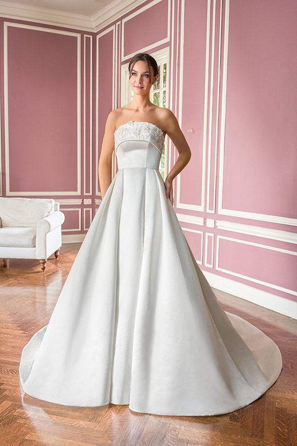 Bride in a strapless satin ball gown wedding dress featuring a delicate floral appliqué neckline, structured bodice, and full skirt, photographed in a romantic blush pink bridal suite.