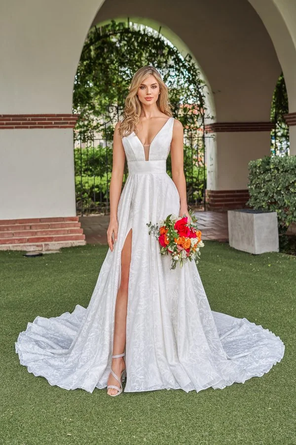 Front view of a jacquard A-line wedding dress featuring a deep plunge neckline with an illusion inset, high leg slit, wide waistband, and sweeping train, paired with a colorful bridal bouquet for a modern bridal look.
