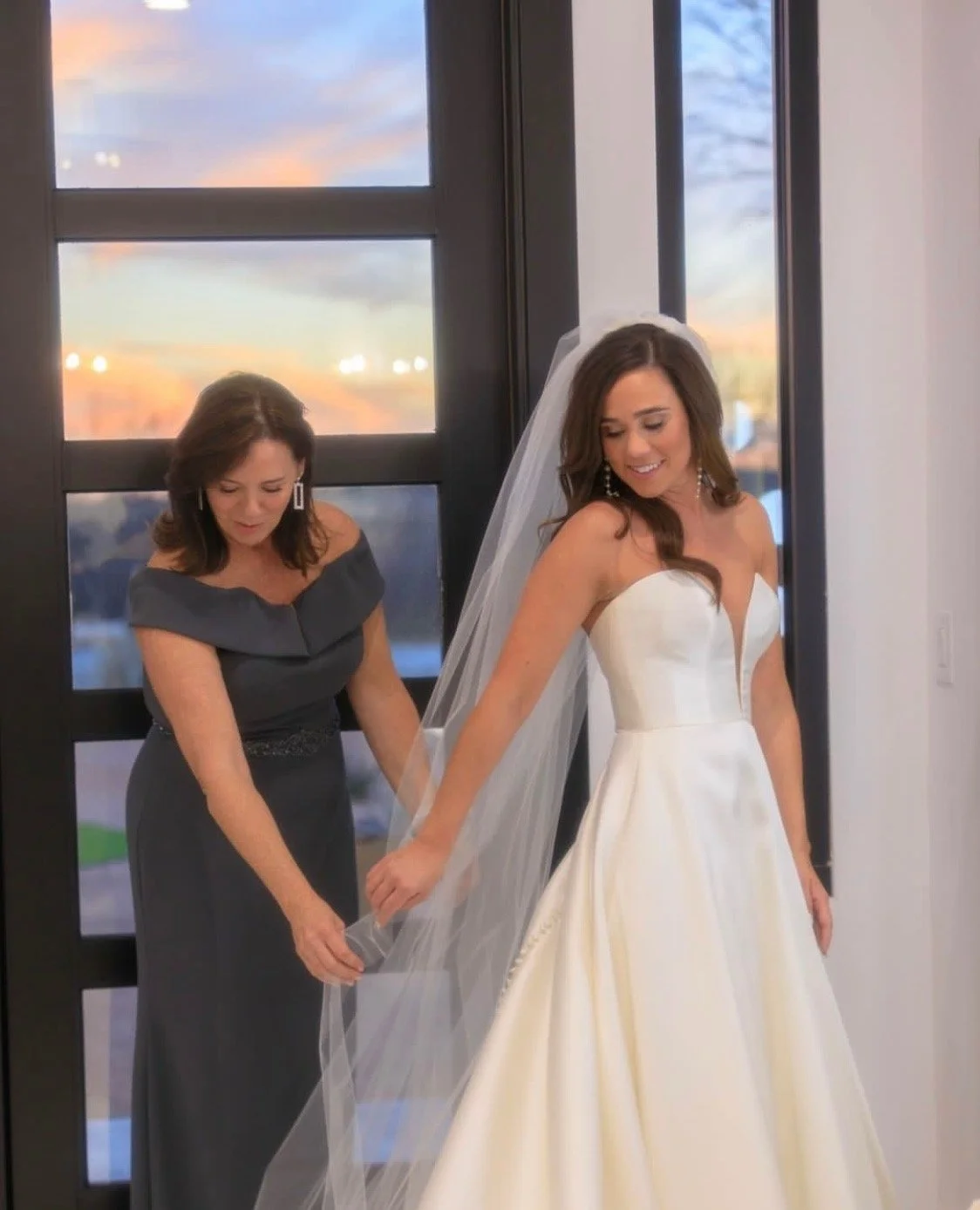 A mother in a charcoal off-the-shoulder gown adjusts her daughter’s veil as the bride smiles in a classic satin strapless wedding dress. They stand by large windows at sunset, sharing a quiet, elegant moment before the ceremony.