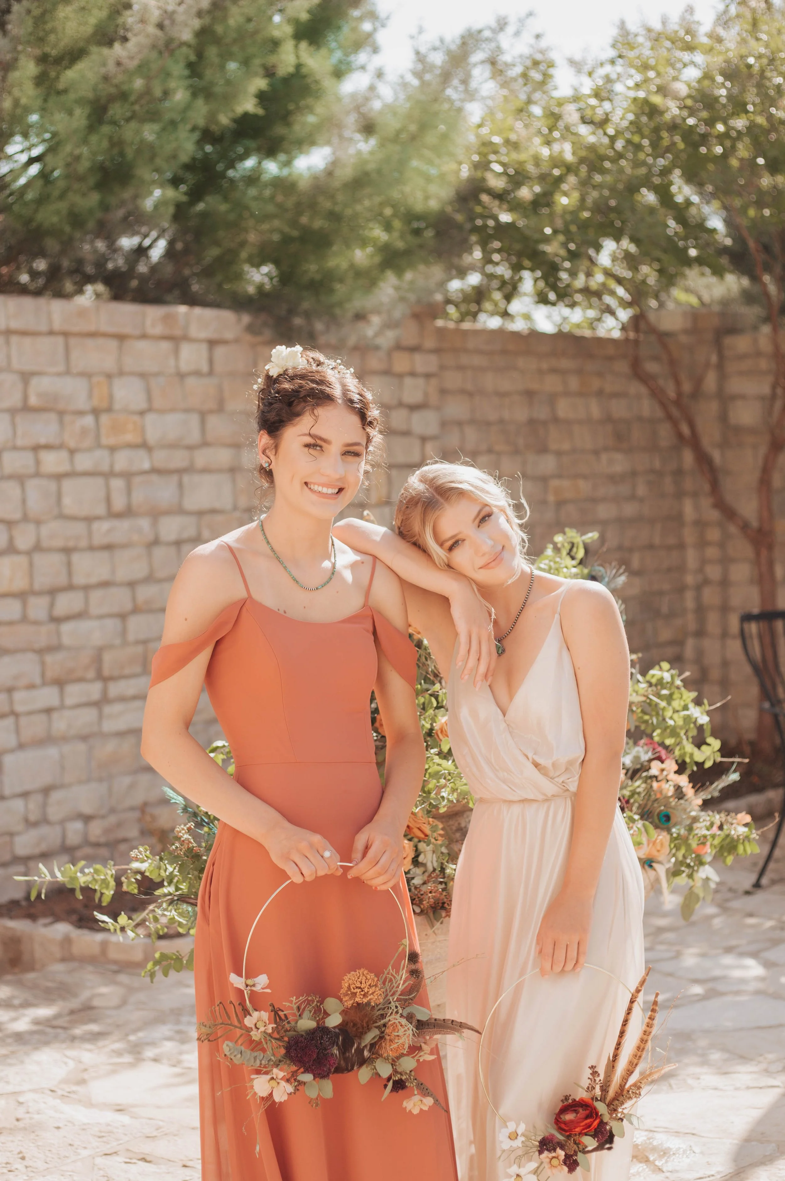 Two women wearing terracotta and soft neutral formal dresses pose with floral hoop bouquets in a sunlit garden setting, showcasing romantic boho-inspired bridal party style and wedding fashion.