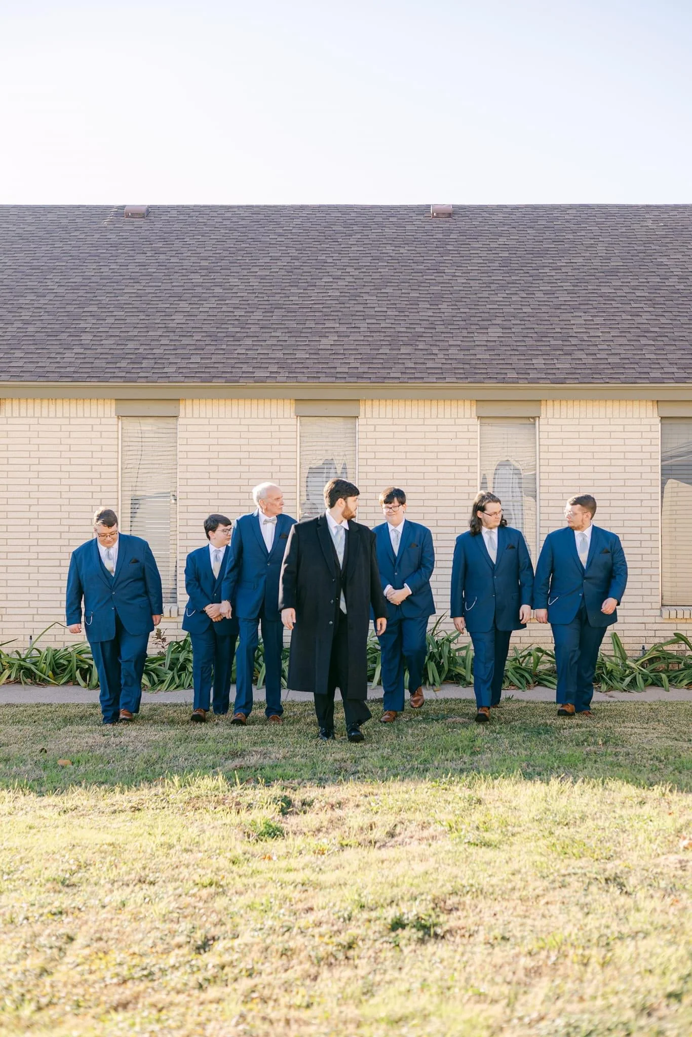 Groom and groomsmen in matching blue suits and formal attire walk together across a lawn outside a wedding venue, capturing a stylish and candid wedding party moment with modern menswear inspiration.