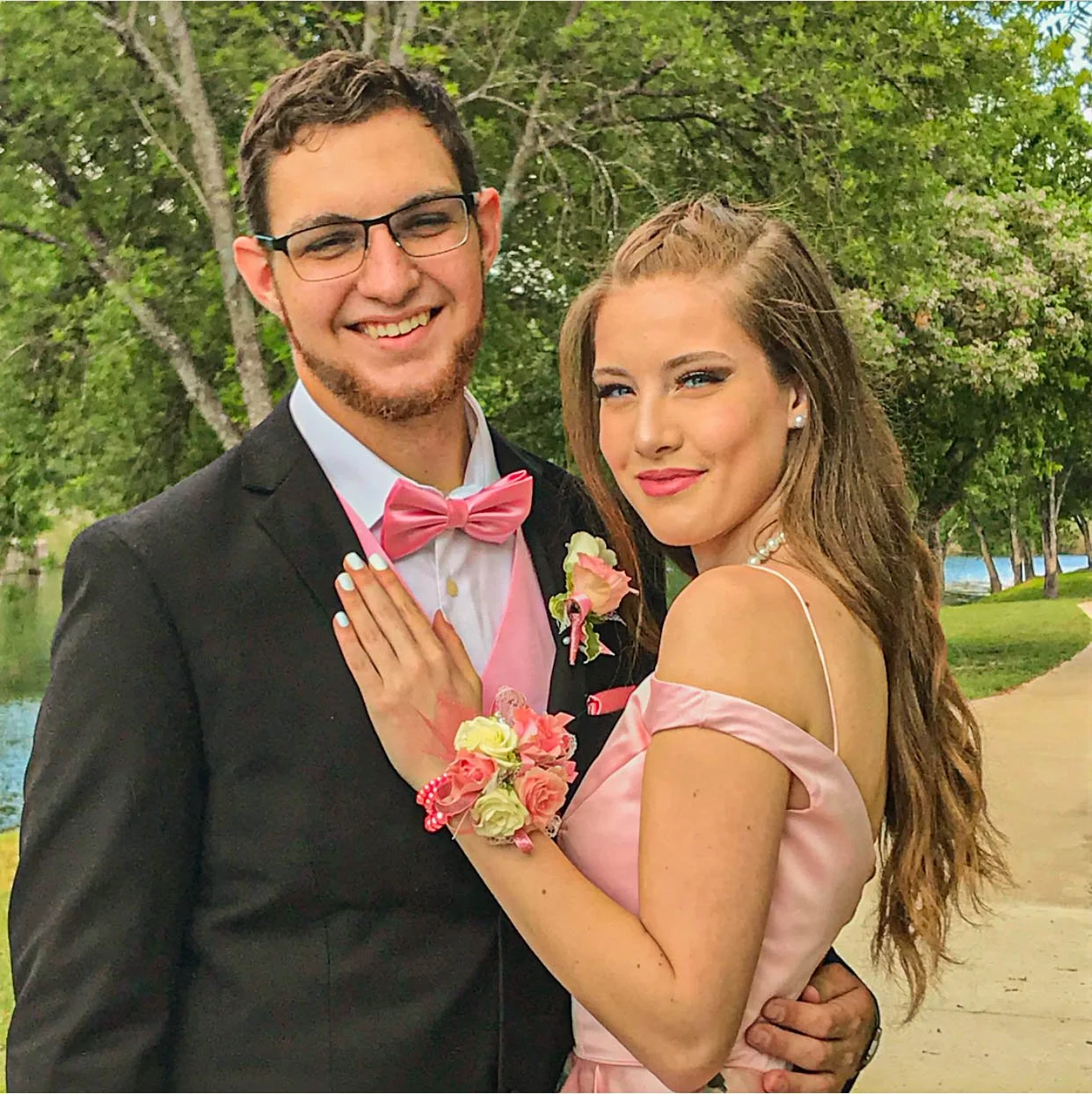 Prom couple in coordinated blush pink and black formalwear pose by the water, featuring a satin off-the-shoulder gown, pink bow tie, and matching floral corsage and boutonniere for a classic and romantic prom portrait.