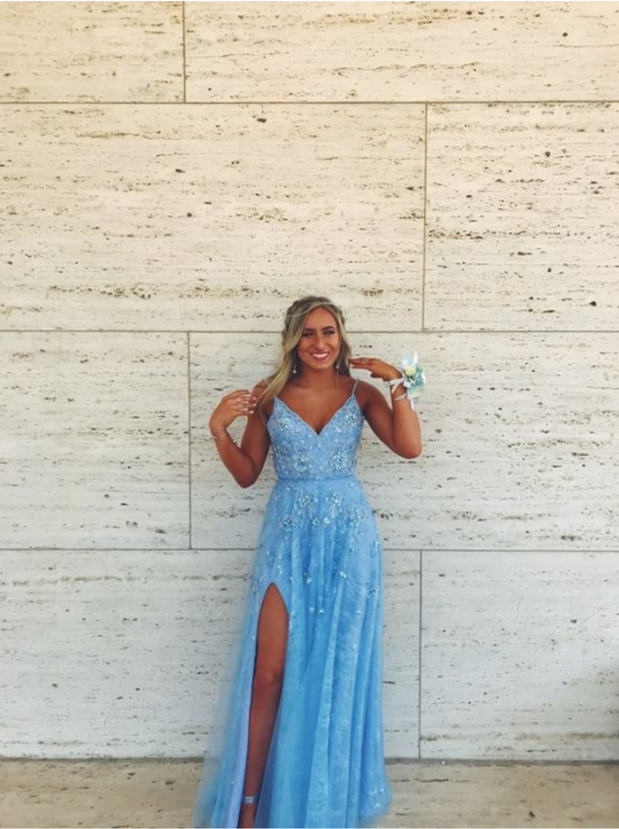 A smiling young woman poses against a wall in a shimmering light blue prom gown featuring intricate beading, a fitted bodice, and a thigh-high slit. The elegant formal look is completed with a coordinating wrist corsage and soft glamour styling.