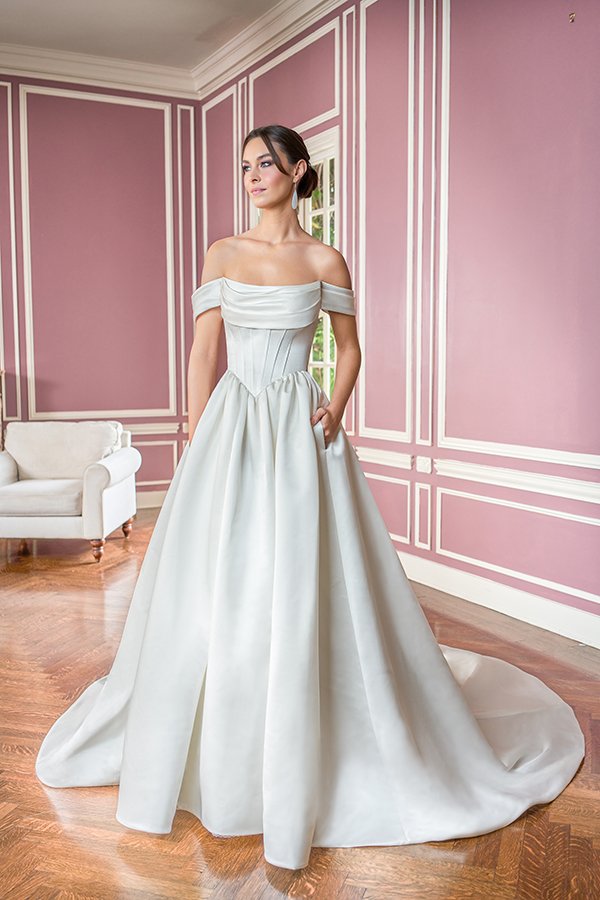 Bride in an off-the-shoulder satin ball gown wedding dress featuring a draped neckline, corset-inspired waist, hidden pockets, and a flowing train, photographed in a romantic blush pink bridal suite.