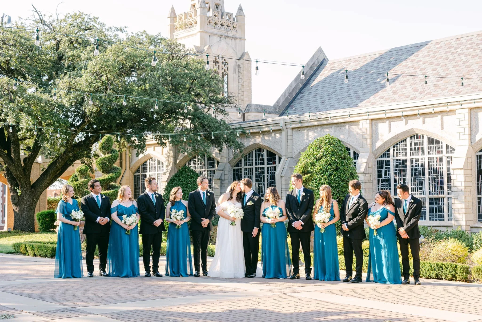 Bride and groom share a romantic kiss surrounded by bridesmaids in teal gowns and groomsmen in black suits, creating a timeless wedding party portrait in front of an elegant stone venue.