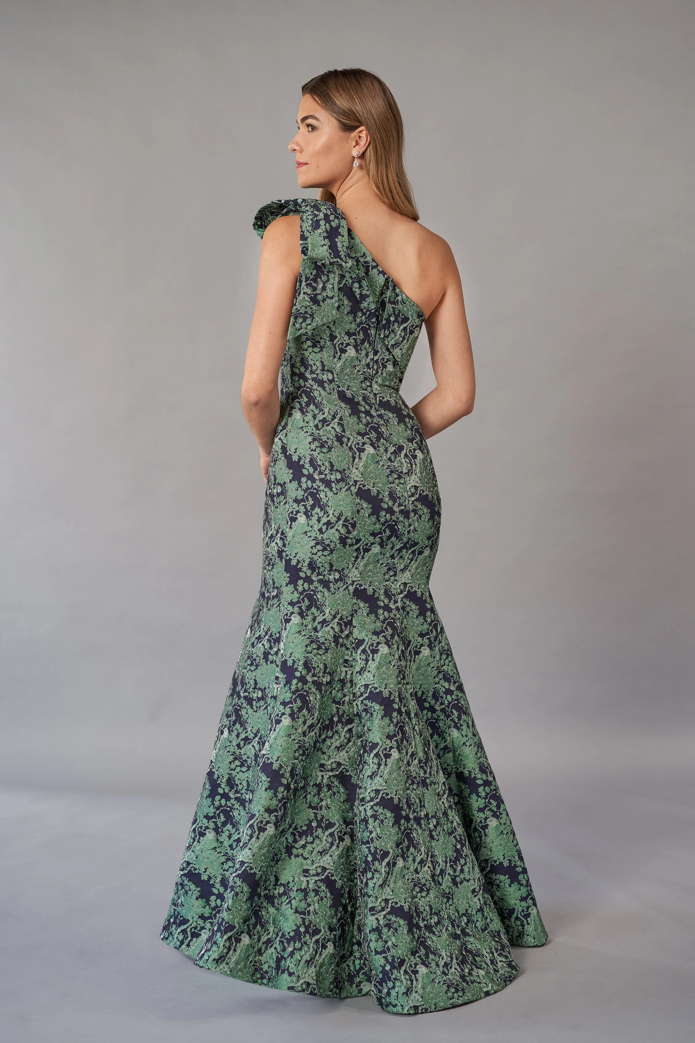 Back view of an elegant green and navy mother of the bride gown with a one-shoulder neckline, jacquard fabric, statement shoulder bow, asymmetrical back, and flattering mermaid silhouette for formal occasions.