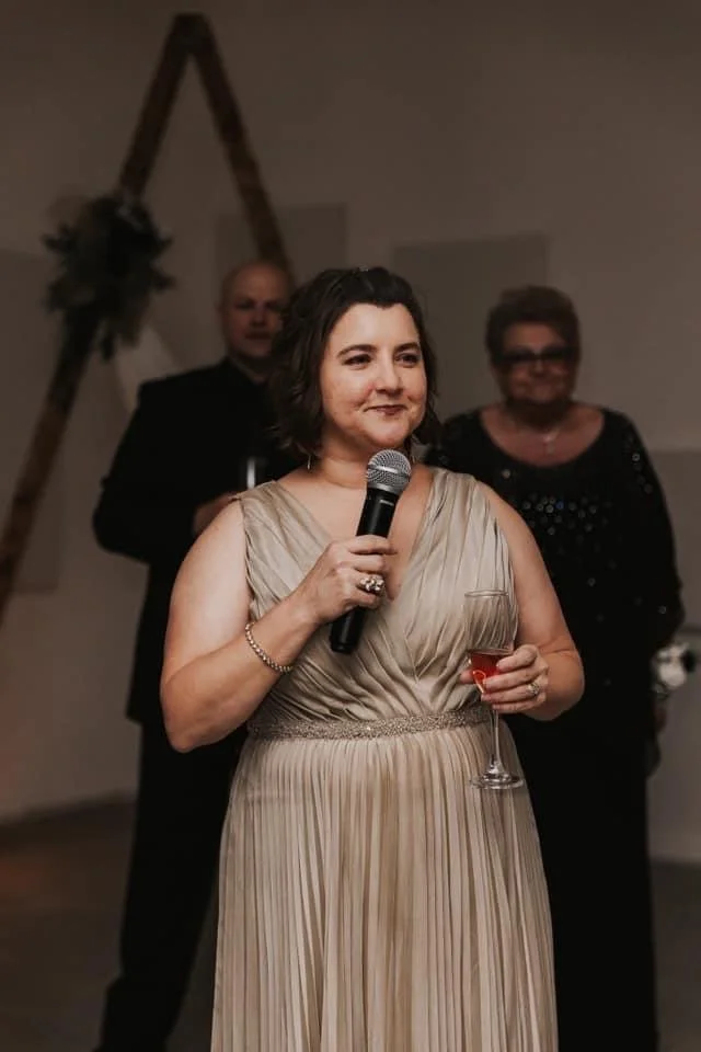 A mother of the groom in a champagne-colored pleated gown holds a microphone and a glass of champagne while giving a speech at a wedding reception. Guests stand behind her as she delivers the toast.