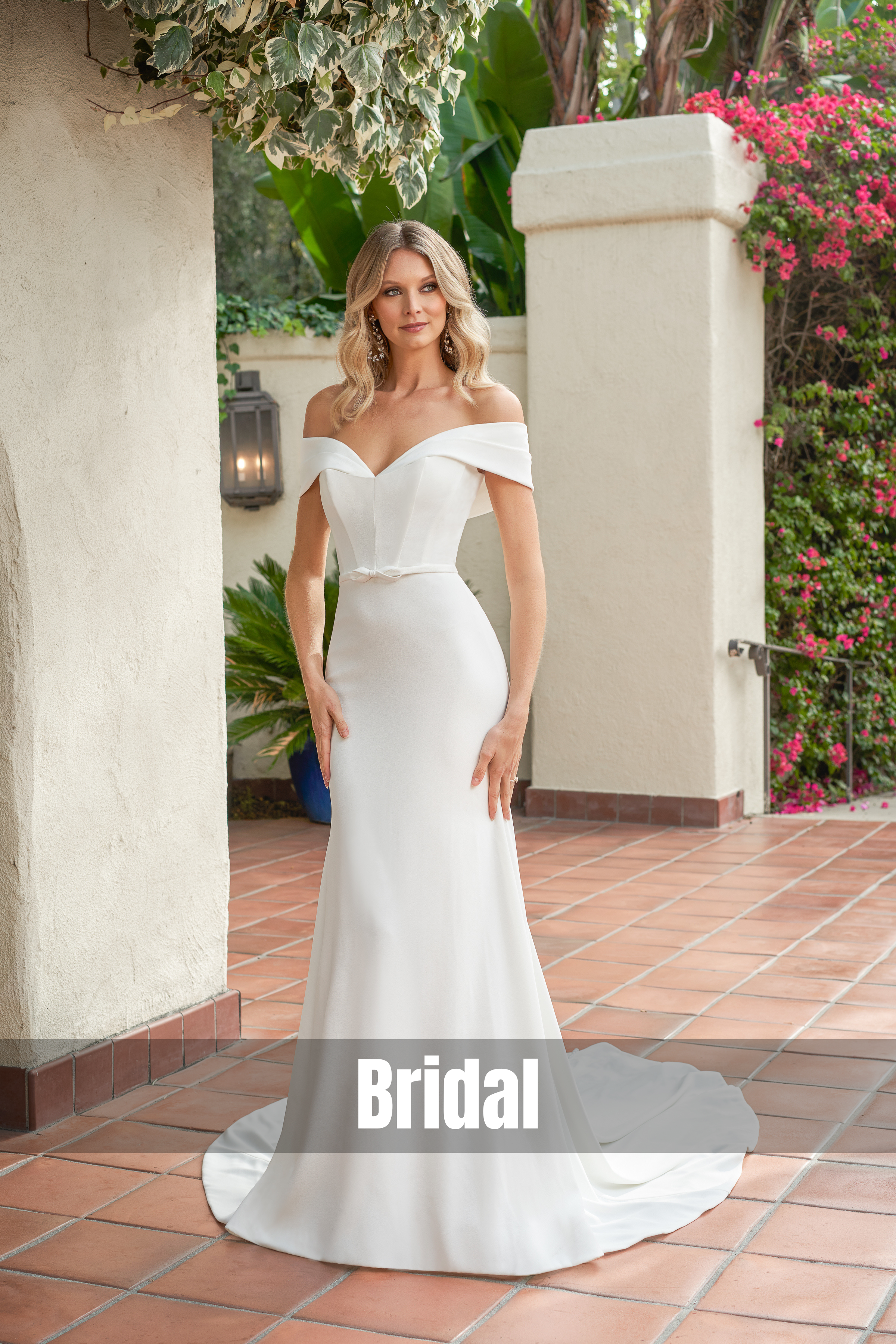 Model wearing an elegant off-the-shoulder white bridal gown with a fitted silhouette and flowing train, posed in a Mediterranean-style courtyard surrounded by greenery and vibrant pink flowers.