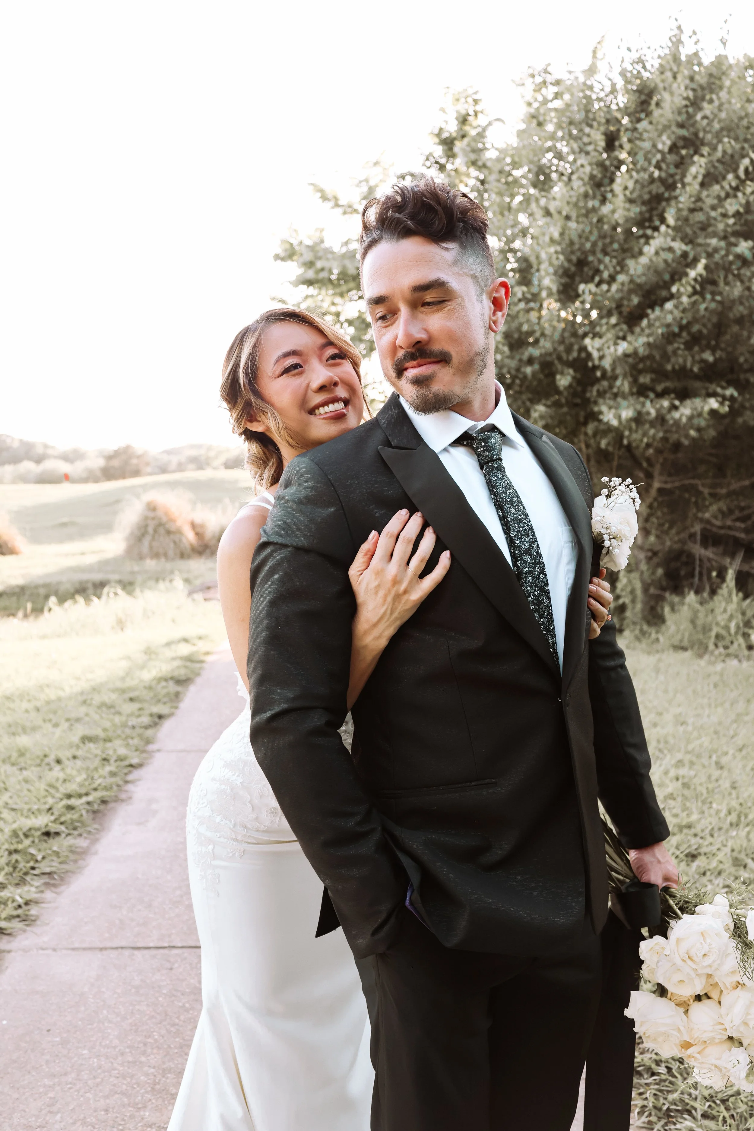 Bride in a sleek white wedding gown wraps her arms around her groom in a black suit, creating a romantic outdoor wedding portrait surrounded by natural scenery and soft golden light.