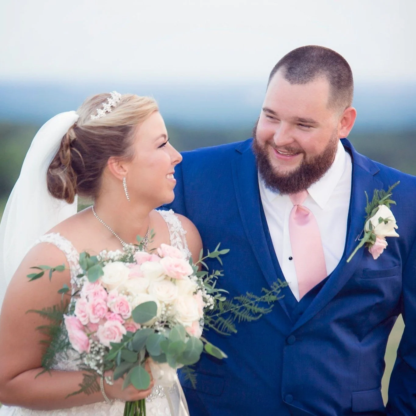 Bride in a lace wedding gown and veil shares a joyful glance with her groom in a royal blue suit and pink tie, holding a bouquet of blush and white roses for a timeless wedding portrait.