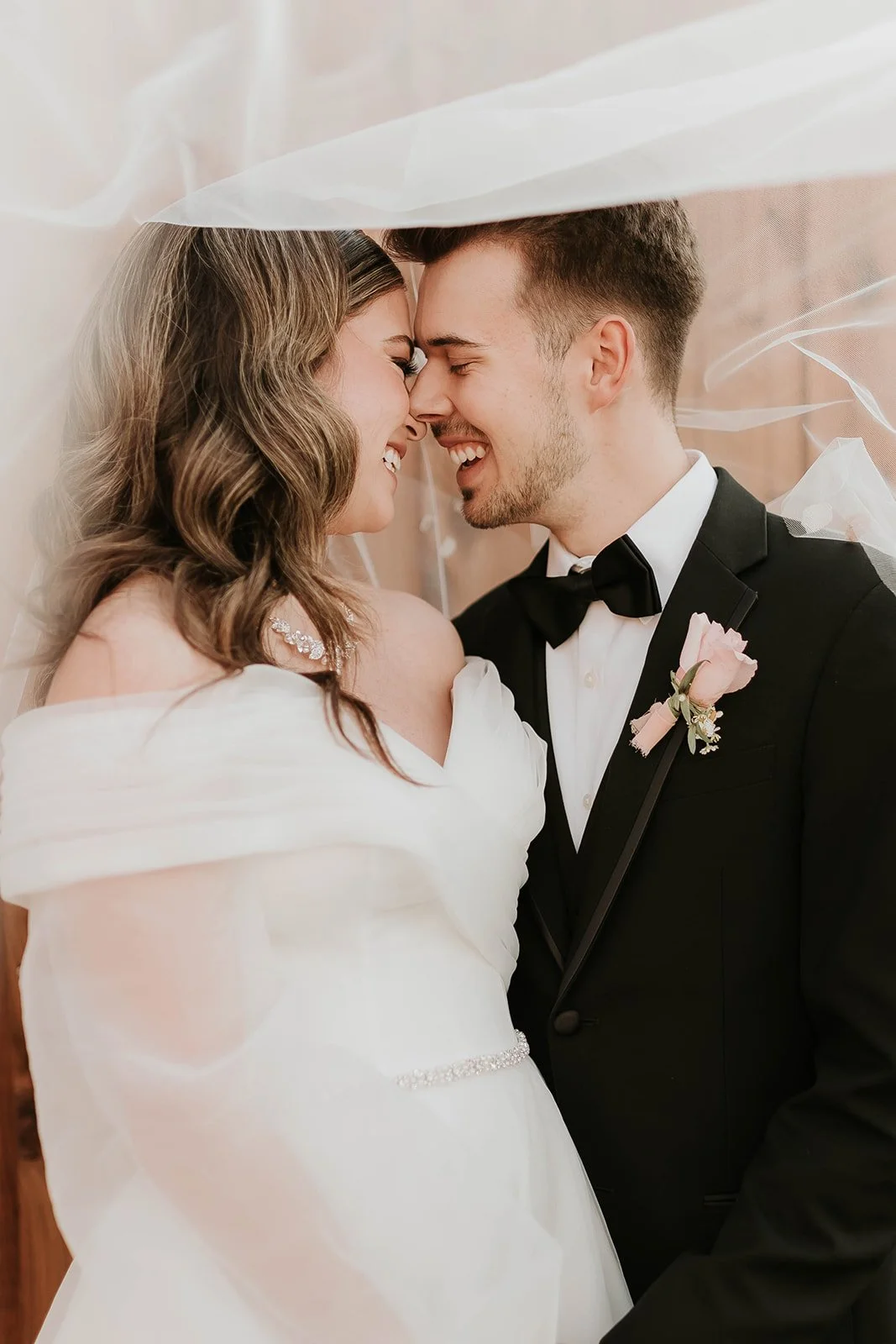 Bride in an elegant off-the-shoulder wedding gown and groom in a classic black tuxedo share a joyful close-up beneath a soft veil, capturing a romantic and timeless newlywed portrait.