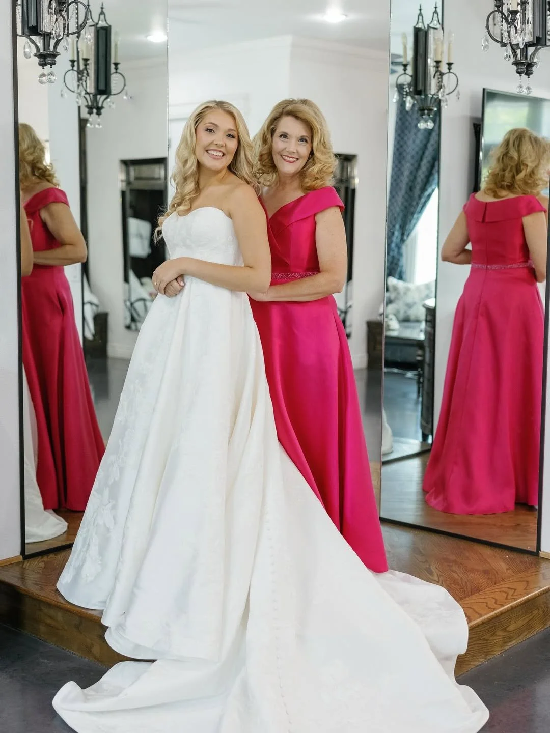 Bride in a classic strapless wedding gown poses with her mother wearing a bright pink off-the-shoulder formal dress, creating a heartfelt mother-of-the-bride moment inside a bridal suite.