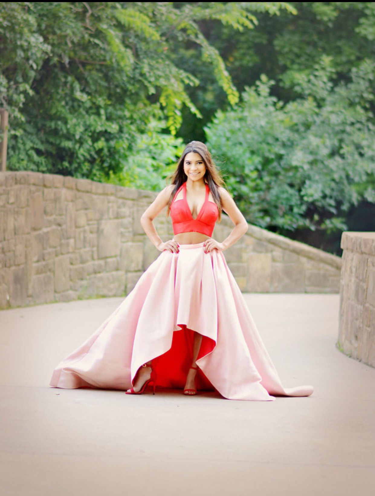 A smiling young woman stands on a stone pathway in a striking two-piece prom dress with a vibrant red halter bodice and flowing blush pink high-low skirt. The outdoor setting and dramatic silhouette create a stylish formal portrait.