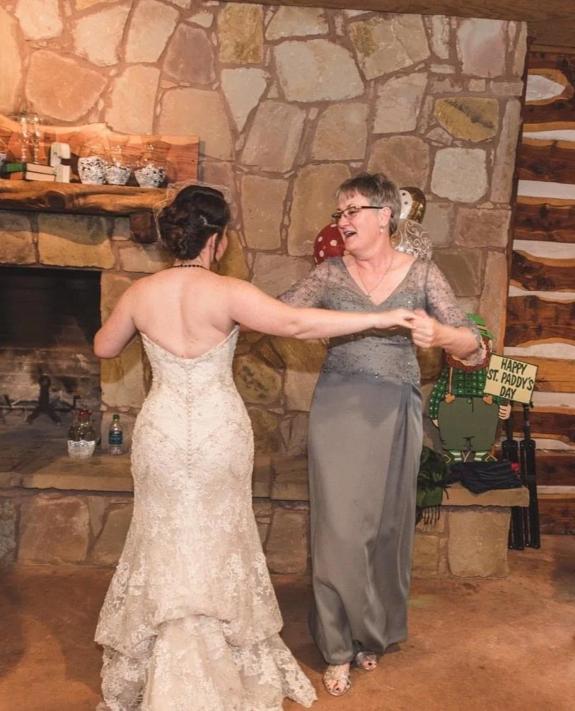 Bride in a lace strapless gown dances with the mother of the bride in a gray dress inside a rustic stone-and-wood venue. Guests smile and clap around them near a fireplace decorated with flowers and St. Patrick’s Day accents.
