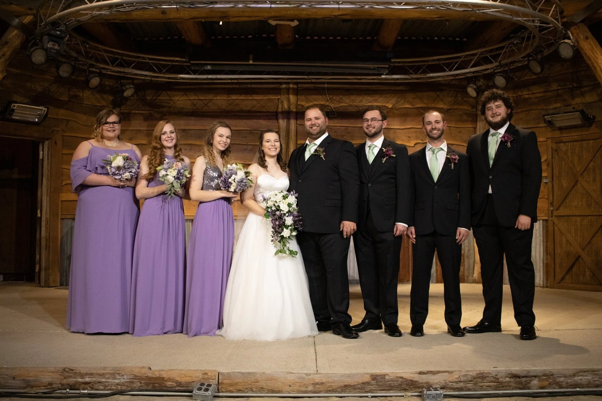 Bride and groom pose with their bridal party on a rustic barn stage, featuring bridesmaids in coordinated lavender gowns, groomsmen in black suits, and elegant floral bouquets for a timeless wedding portrait.