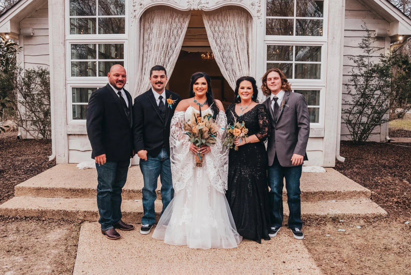Bride in a lace off-the-shoulder gown poses with her family outside a white chapel. Her mother wears a black beaded gown, and the men wear jackets with jeans for a rustic, elegant wedding look.