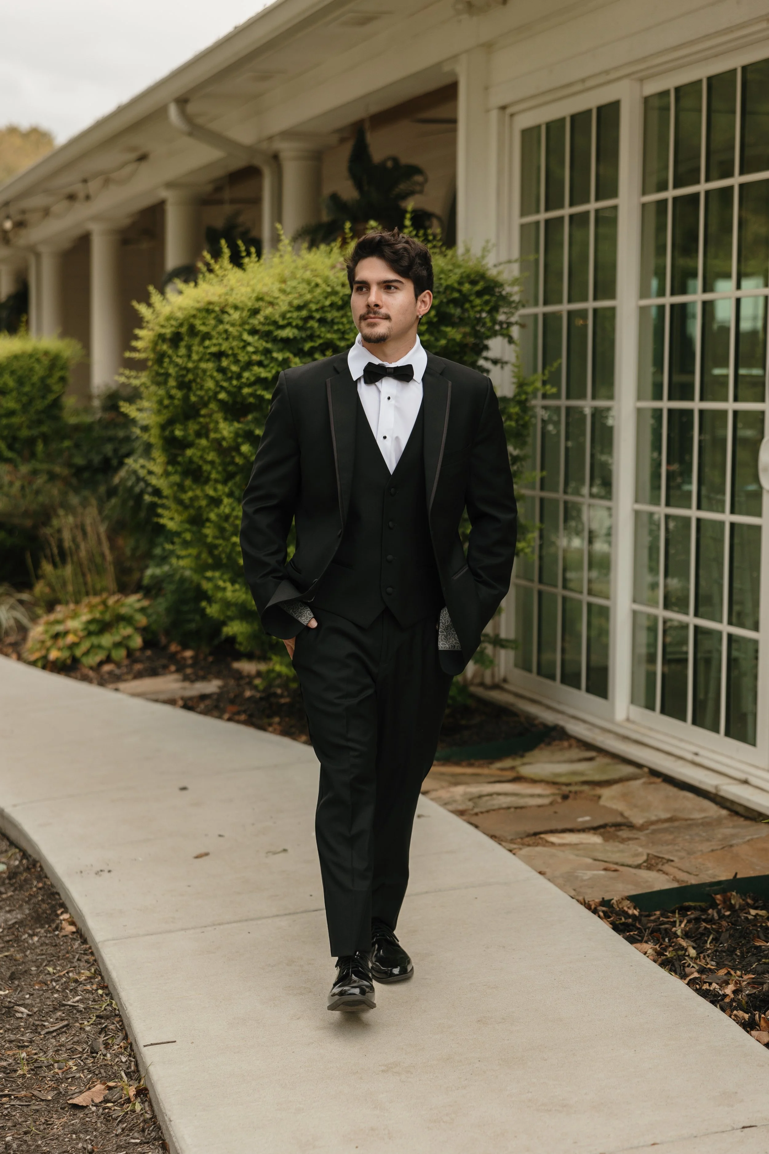 Model showcasing a classic black tuxedo with a matching vest and bow tie, walking outside a sophisticated wedding venue, highlighting timeless groom style and formal wedding menswear.
