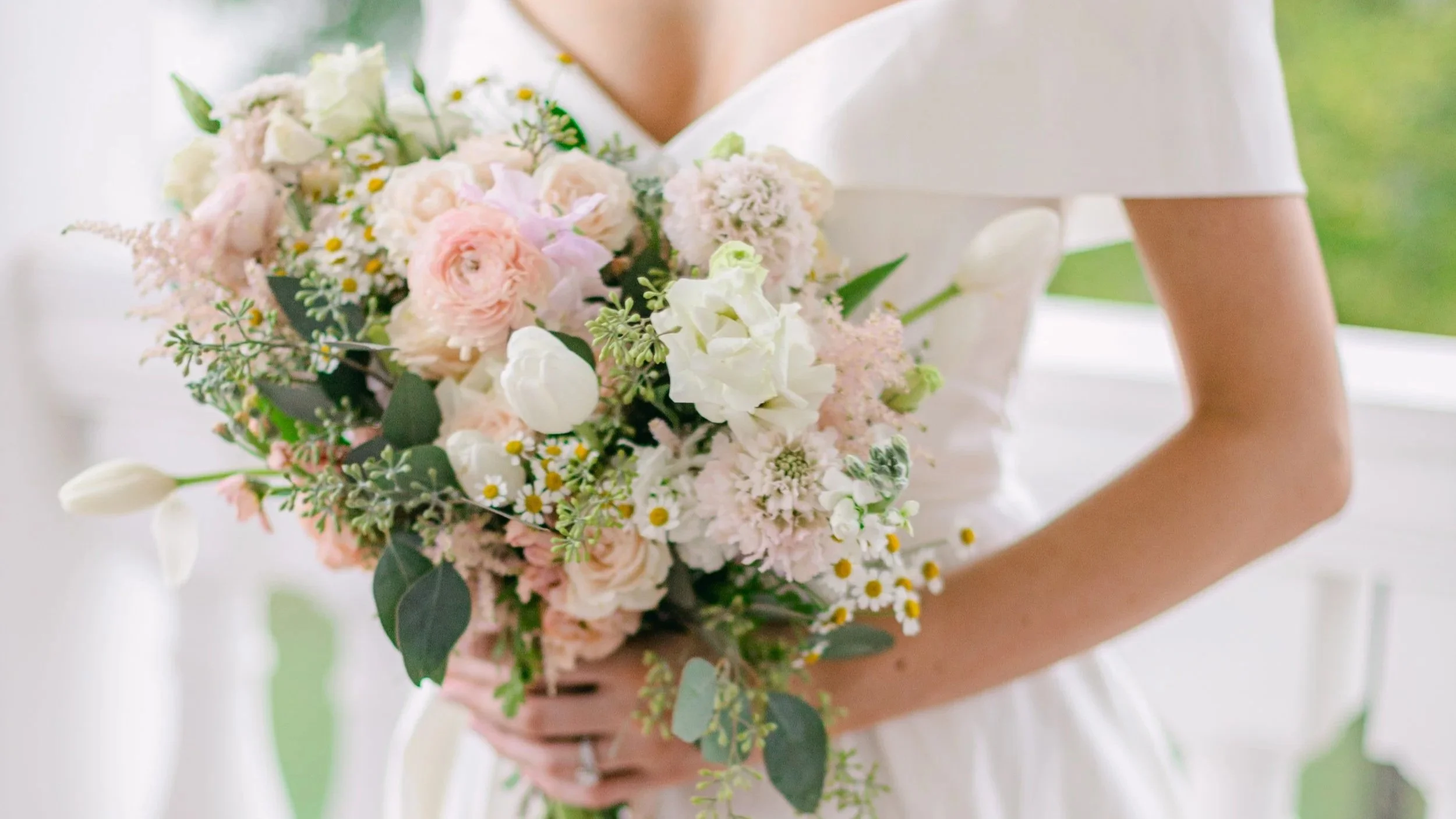 Close-up of a bride in an off-the-shoulder wedding gown holding a romantic blush and white bridal bouquet with roses, tulips, daisies, and greenery, showcasing elegant wedding flowers and timeless bridal style.