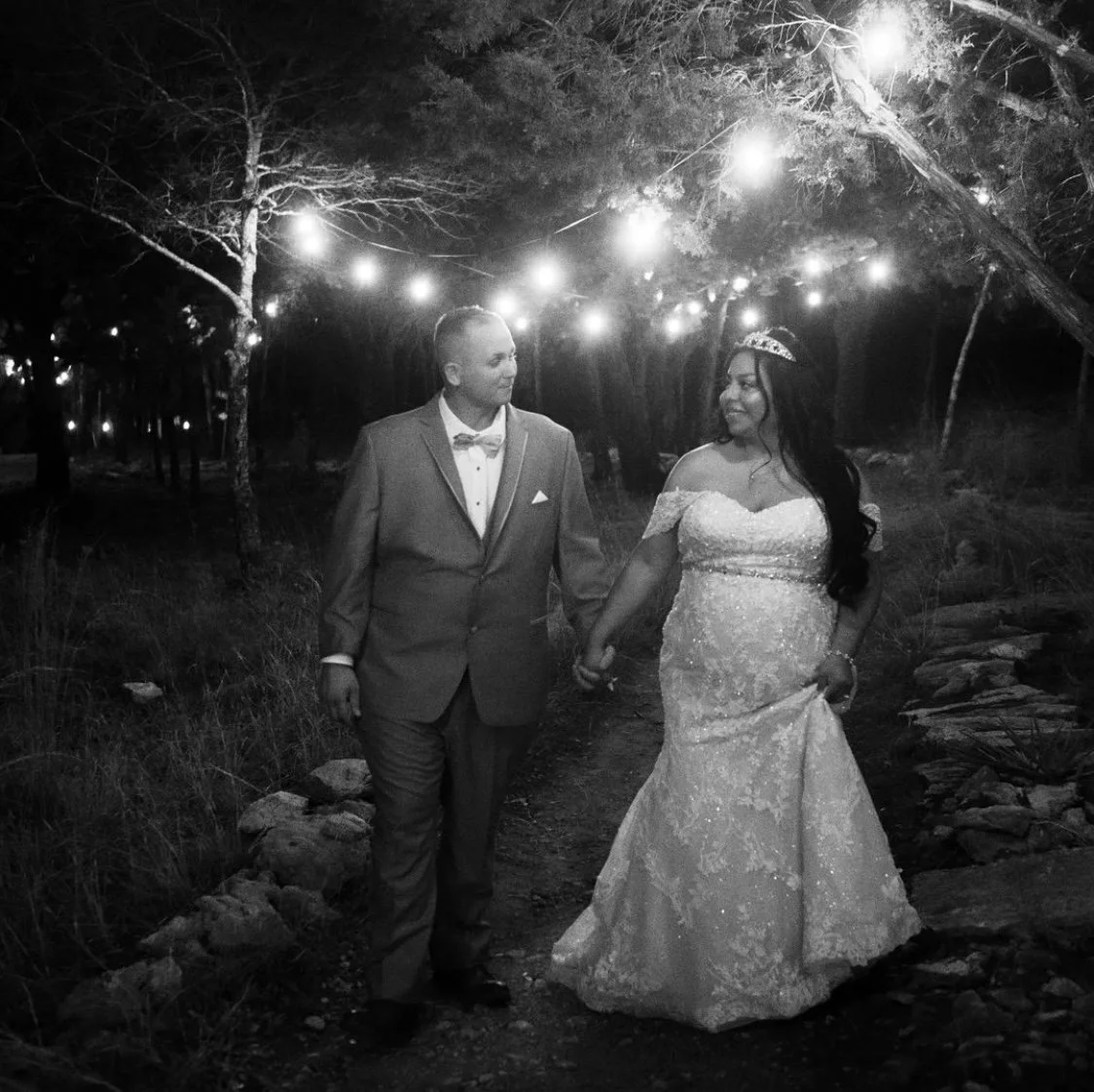Bride in a sparkling lace wedding gown and tiara walks hand in hand with her groom beneath twinkling string lights, creating a romantic nighttime wedding portrait filled with warmth and elegance.