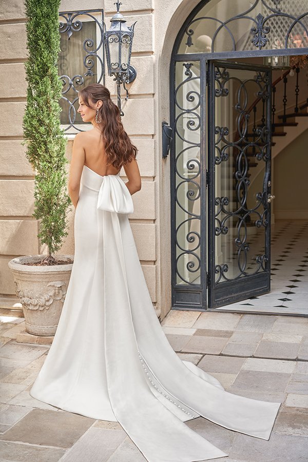 Bride showcases a minimalist satin wedding dress with a statement bow back, open back, and flowing cathedral train at an elegant estate entrance, blending timeless sophistication with modern bridal style.