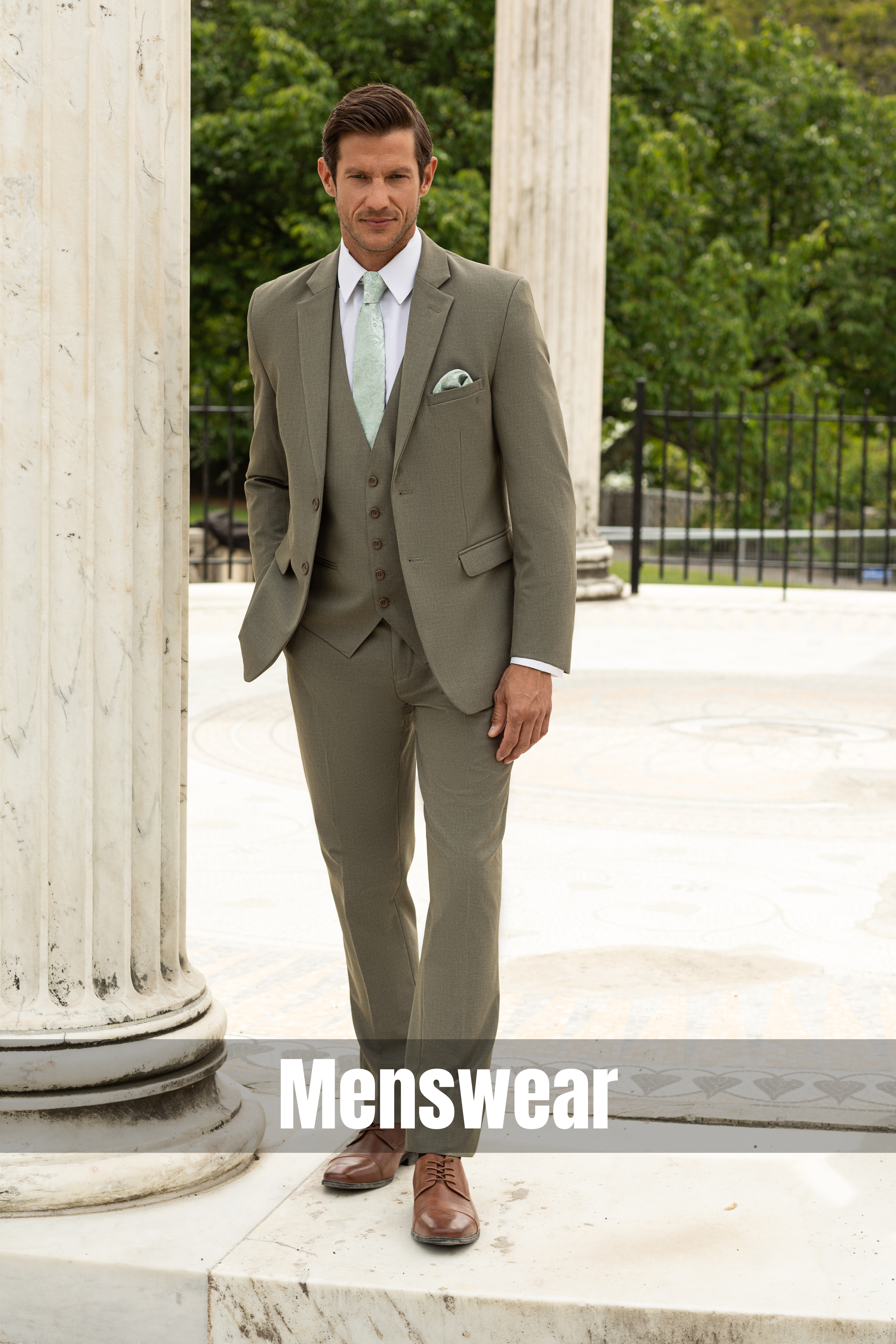 Model wearing a tailored light olive three-piece wedding suit with a coordinating vest, mint tie, and pocket square, posed among classical stone columns in an elegant outdoor venue.