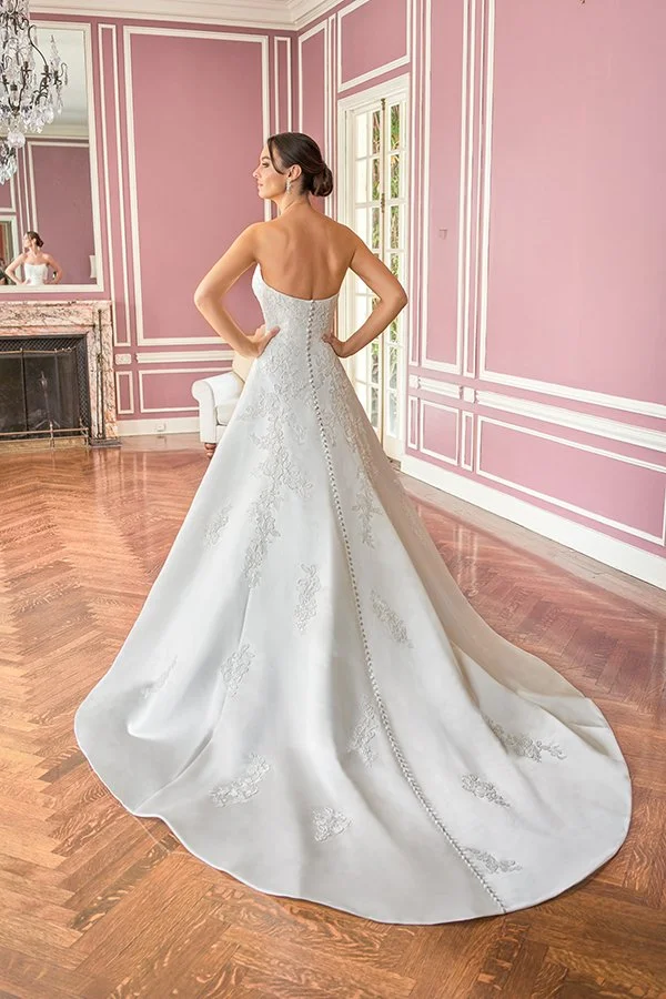 Bride showcasing the back of a square neckline satin A-line wedding dress with floral appliqués, elegant button-back detailing, and a flowing chapel train, photographed in a romantic blush pink bridal suite.