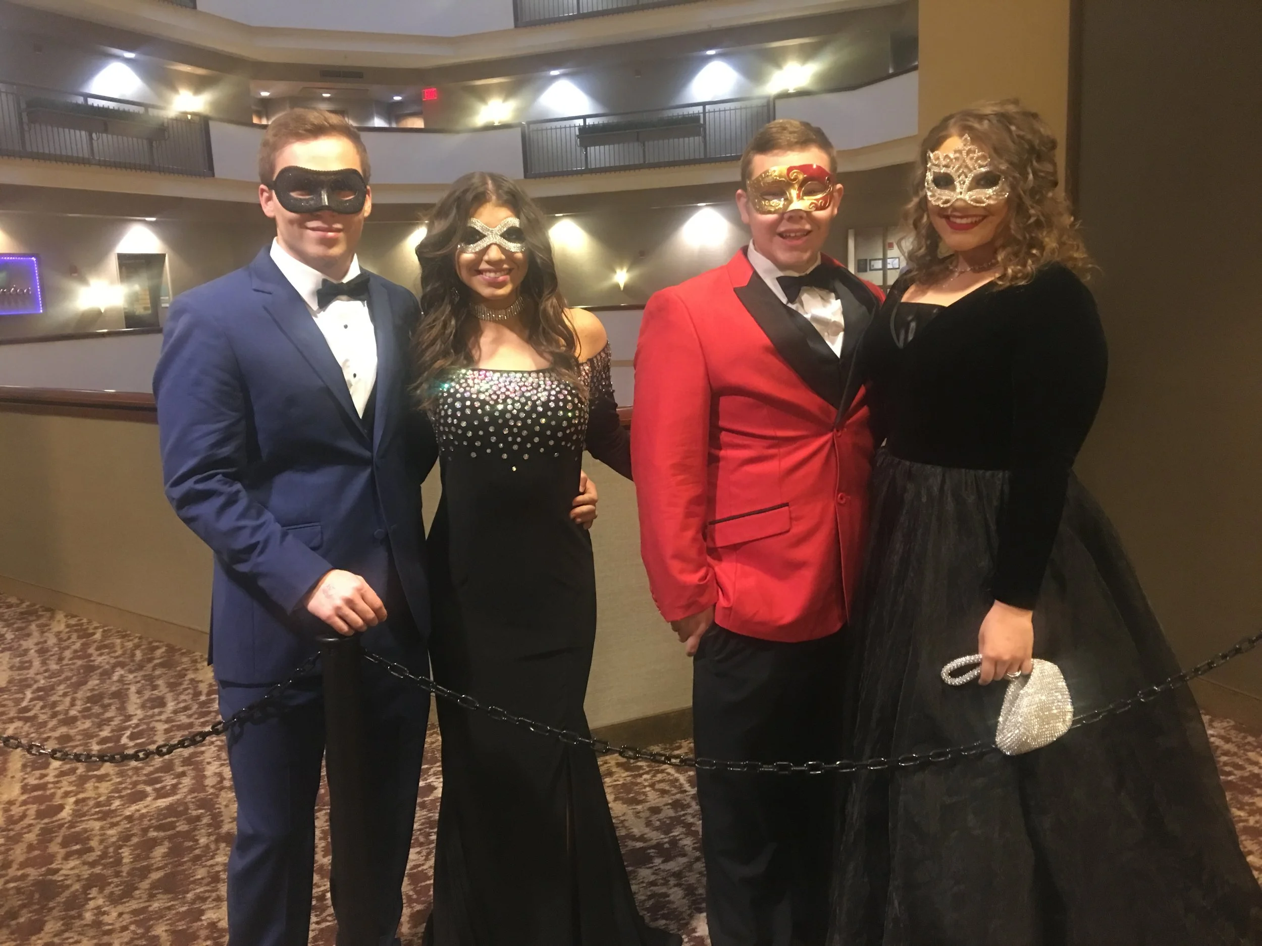 Two  prom couples dressed in sophisticated formalwear and ornate masquerade masks pose at an elegant ballroom venue, showcasing colorful dinner jackets, black evening gowns, and glamorous masquerade-inspired style.