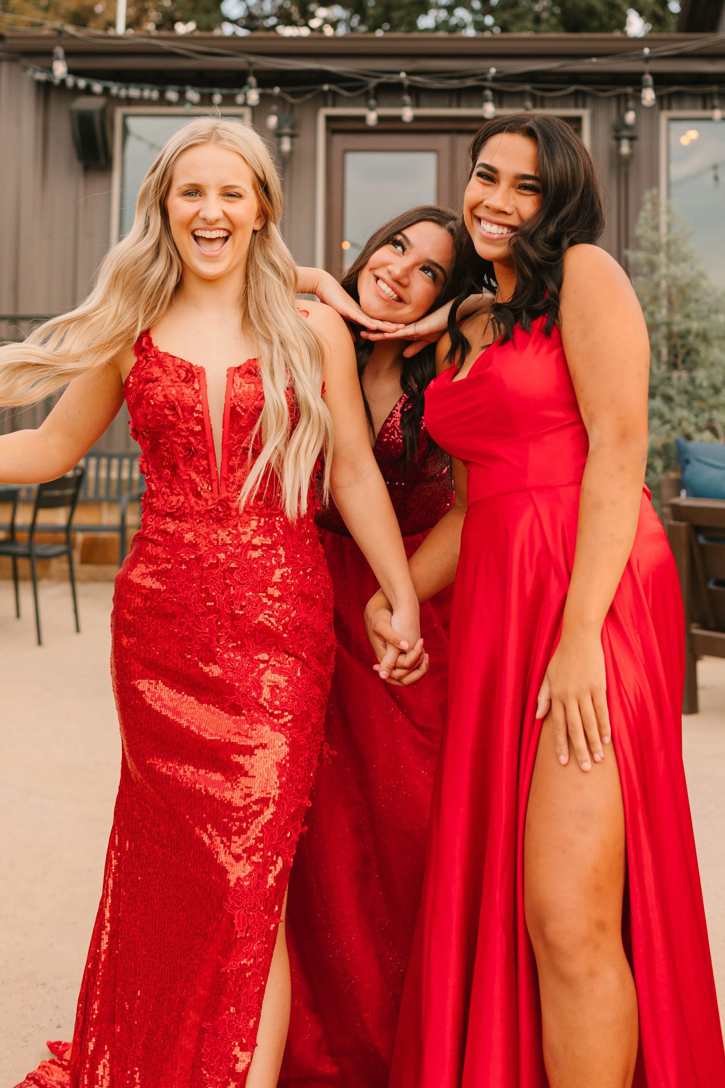 Three friends pose together in striking red formal gowns, smiling and holding hands outside a venue. The image highlights glamorous prom or gala fashion, featuring sequined details, flowing fabrics, and coordinated red eveningwear.