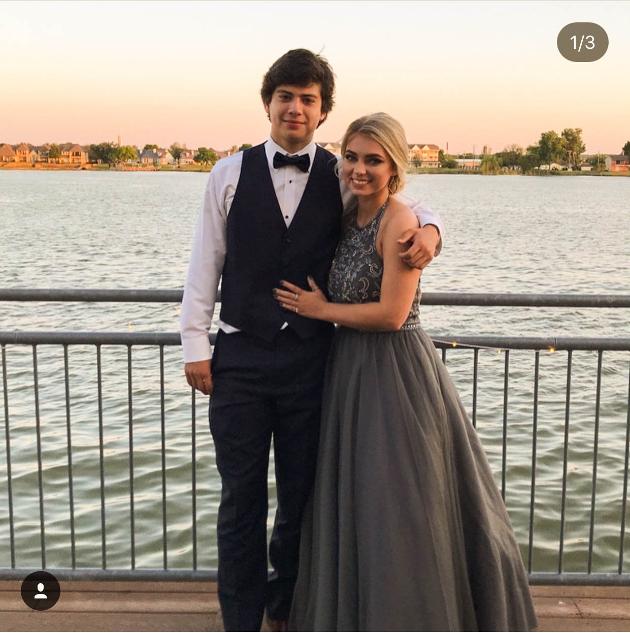 A young couple stands together by a lake at sunset, dressed in elegant formalwear. The young woman wears a charcoal gray gown with intricate beading, while her date wears a classic black vest and bow tie, creating a timeless prom portrait.