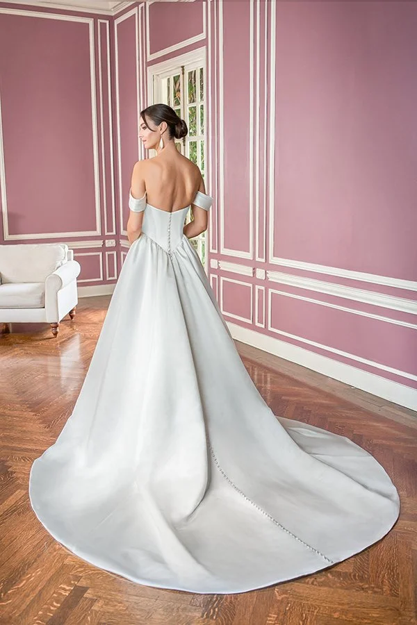 Bride showcasing the back of an off-the-shoulder satin ball gown wedding dress with a button-back bodice and dramatic cathedral train, photographed in a romantic blush pink bridal suite.
