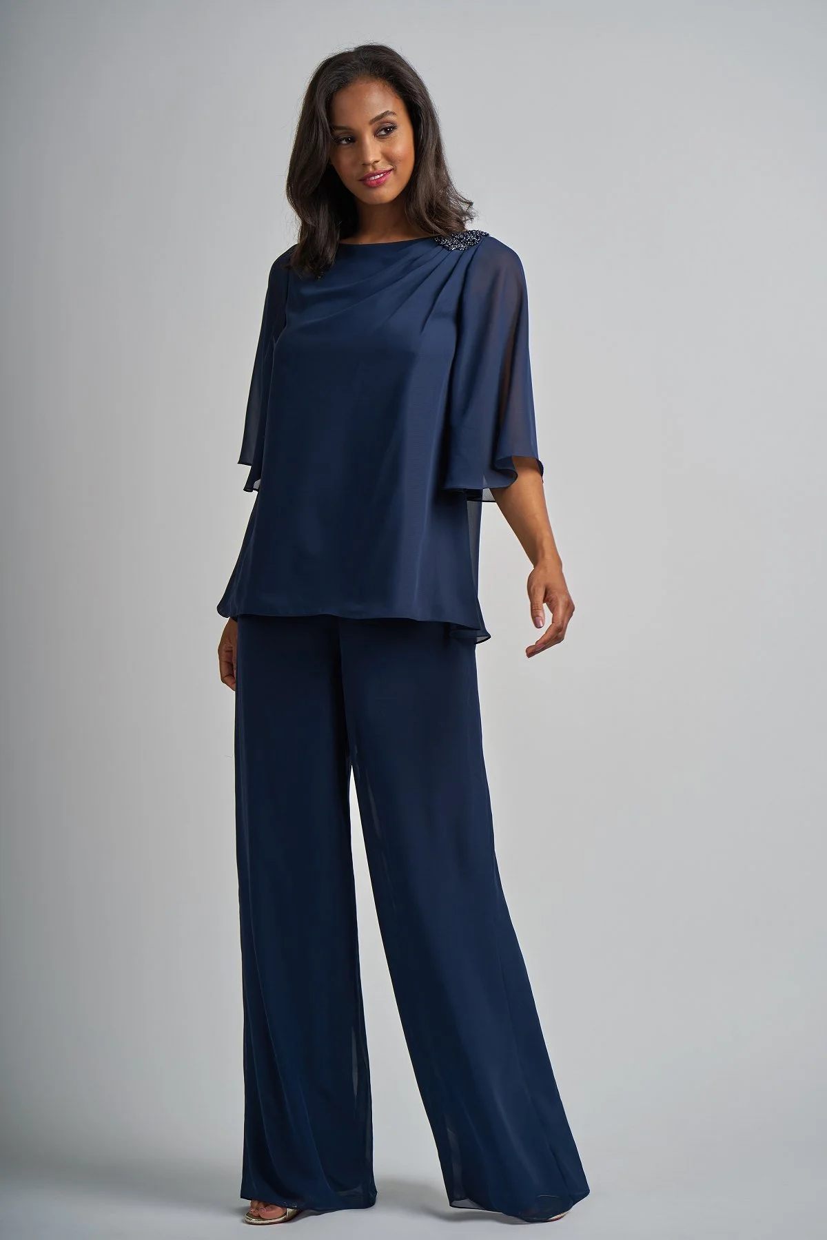 Woman modeling a navy mother of the bride trouser suit featuring wide-leg chiffon trousers, a flowing flutter sleeve top, and elegant beaded shoulder embellishment for special occasions.
