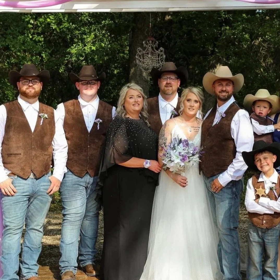 Bride in a white wedding gown poses with family members in coordinated western-inspired attire, including cowboy hats and leather vests, creating a charming country wedding portrait with rustic outdoor appeal.