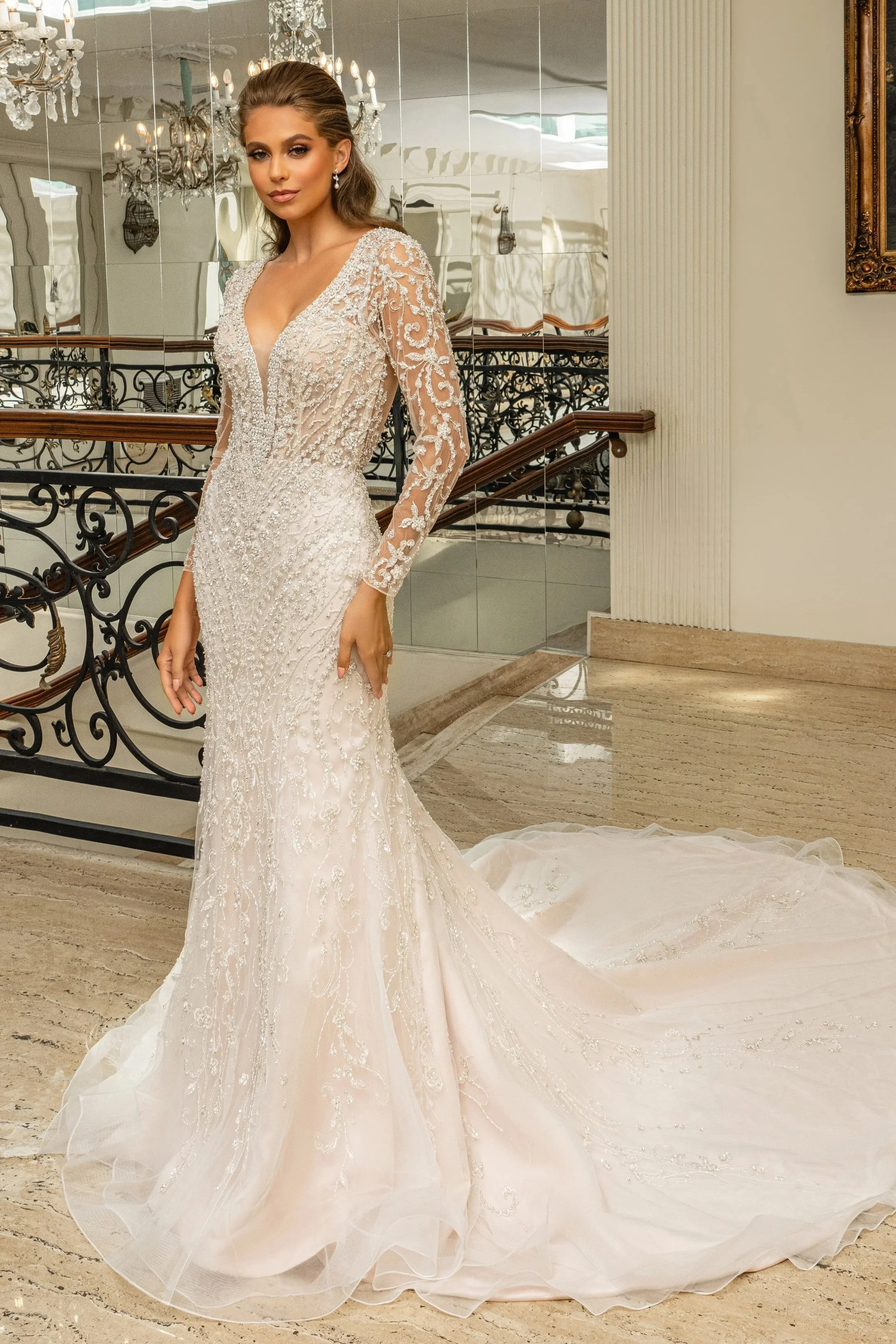 Bride models a long-sleeve beaded mermaid wedding dress with an illusion V-neckline, intricate embellishments, and a flowing tulle train, showcasing elegant bridal fashion and timeless wedding gown inspiration in a luxurious venue.