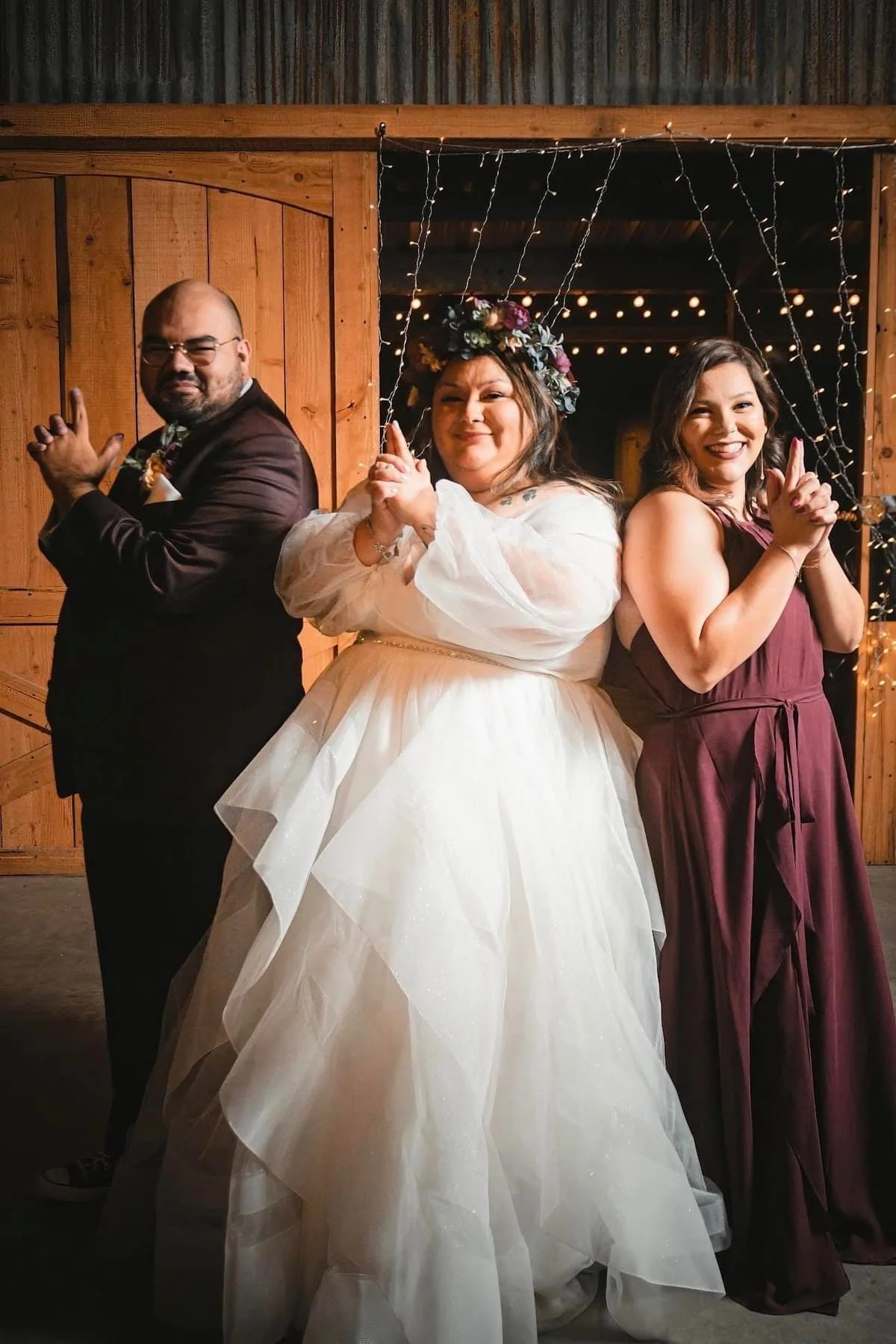 Bride in a romantic white gown poses with two wedding party members in a playful, James Bond-inspired stance, creating a fun and memorable wedding portrait inside a warmly lit rustic barn venue.