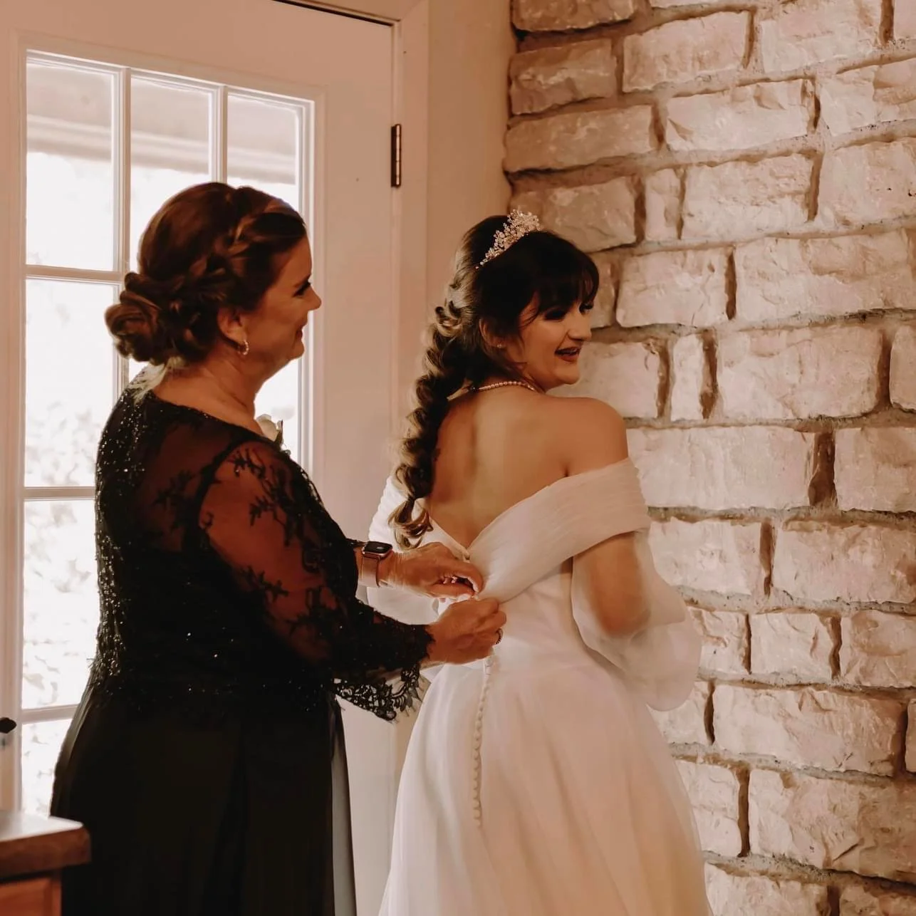 A mother helps fasten her daughter’s off-the-shoulder wedding gown beside a stone wall, both smiling as soft light filters in through a nearby door.