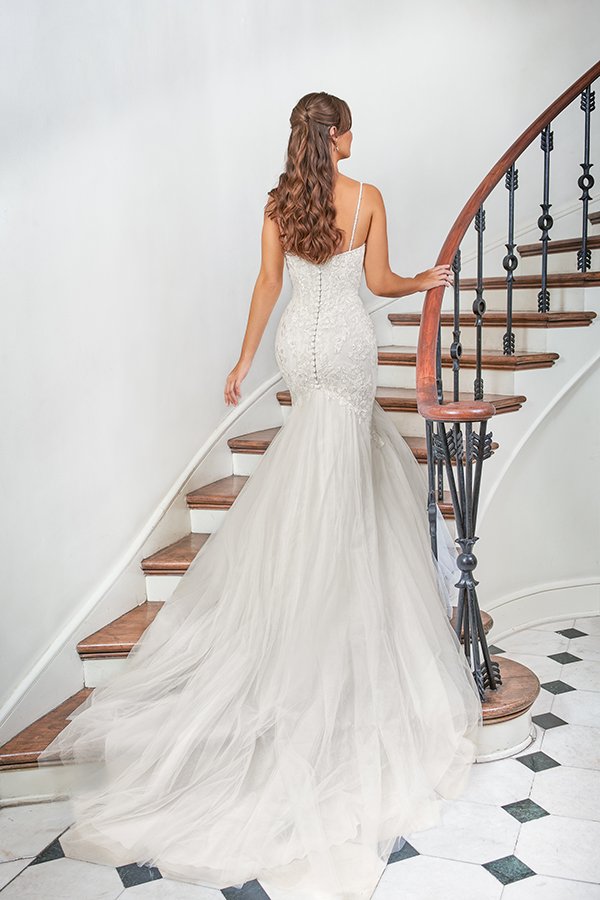 Back view of a bride in a lace mermaid wedding dress with delicate spaghetti straps, covered buttons, and a dramatic tulle train ascending a curved staircase, highlighting timeless elegance and romantic bridal fashion.
