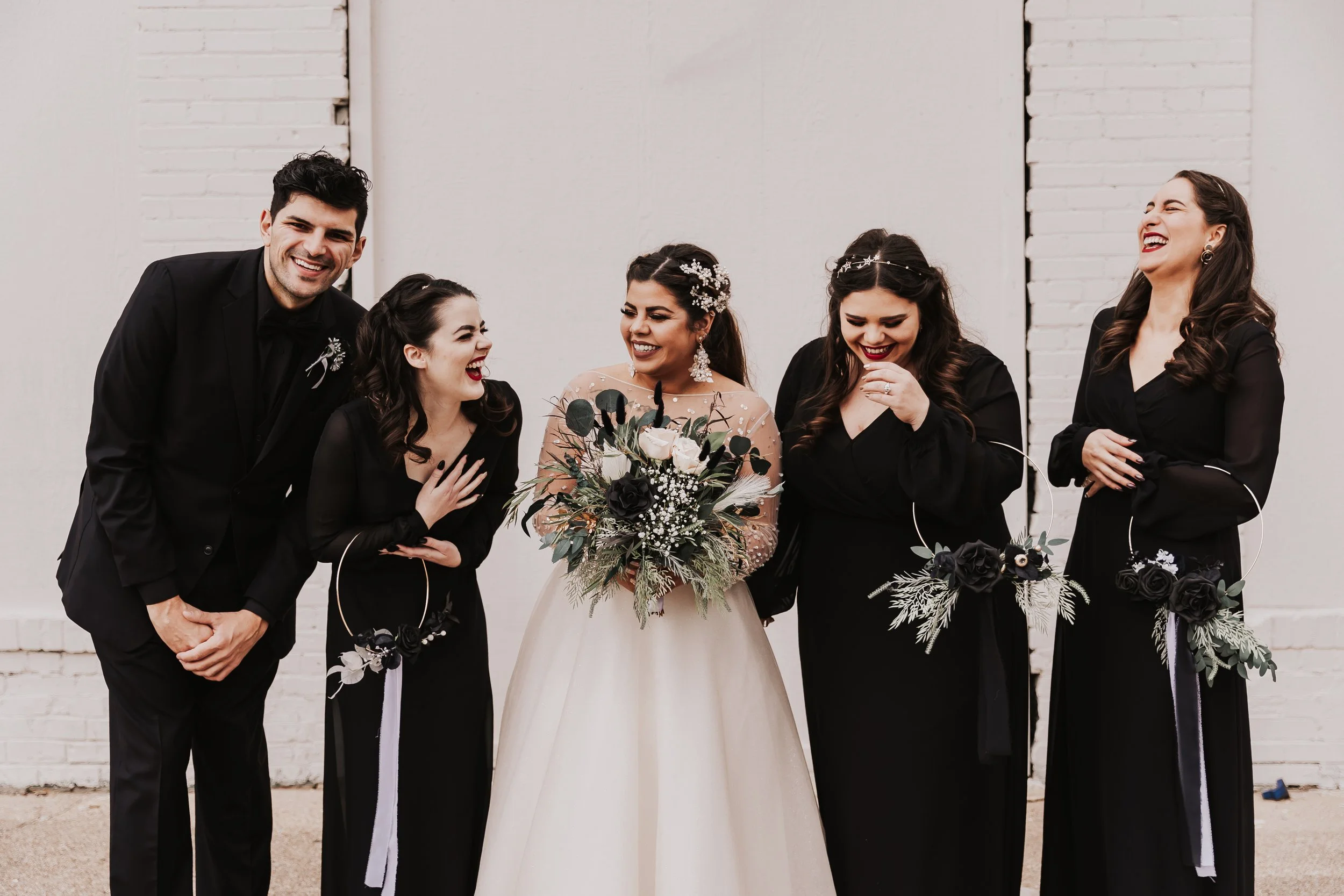 Bride in a romantic white gown shares a joyful moment with her wedding party in coordinated black attire, holding textured bouquets and laughing together for a modern, stylish wedding portrait.
