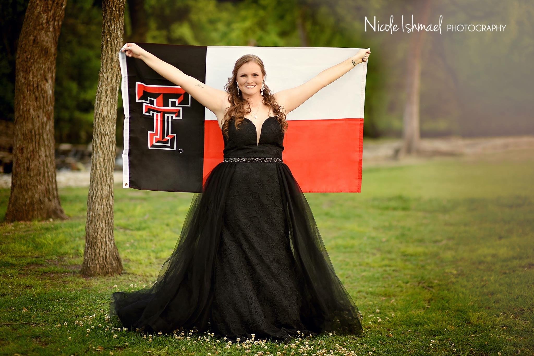 Young woman in a flowing black formal gown stands outdoors on a grassy field, holding a large Texas Tech flag with arms outstretched. Trees and soft natural light create a celebratory portrait setting.