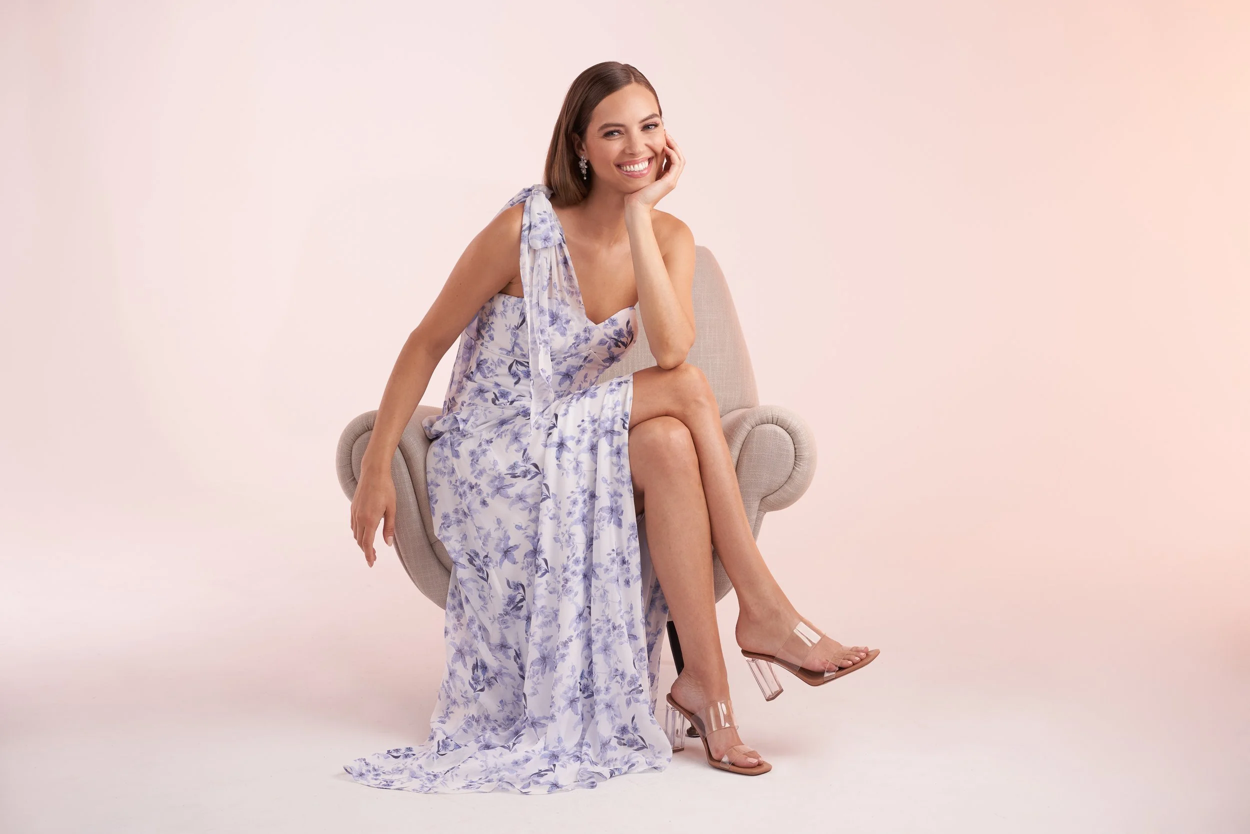 Model seated in a chair wearing a one-shoulder white and blue floral dress, styled with clear heels and delicate earrings, showcasing an elegant bridesmaid look for spring and summer events.