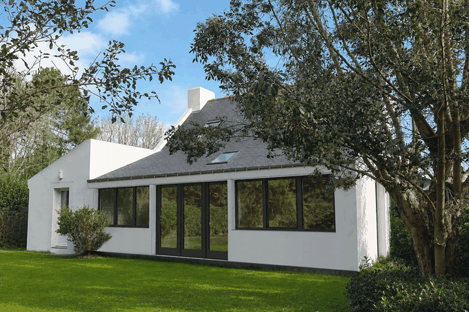 Une maison moderne avec des grandes fenêtres, entourée d'un jardin avec un arbre et des plantes, sous un ciel bleu avec quelques nuages.