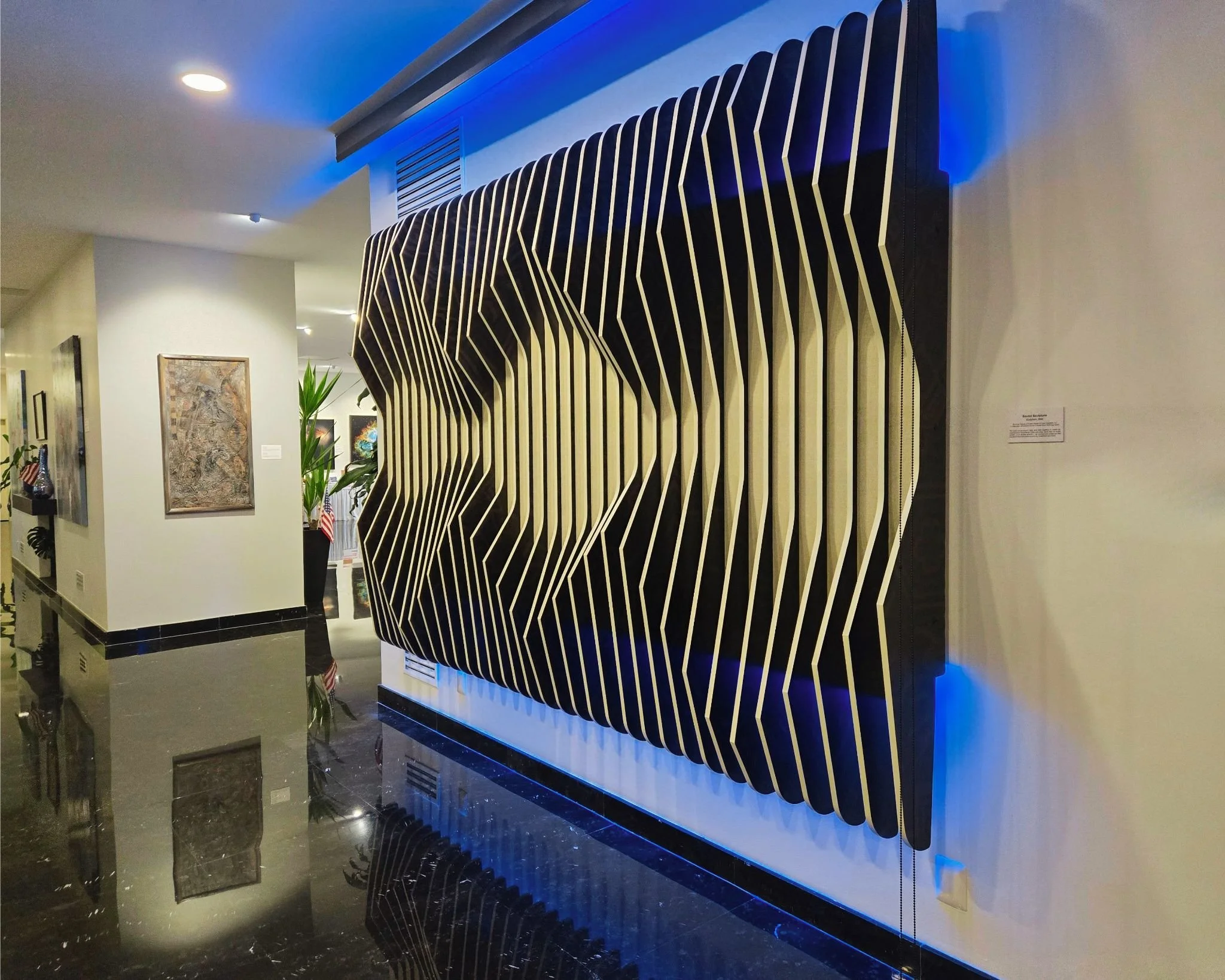 Side-angle view of the completed Sculptura sound sculpture mounted in a modern hall, glowing with integrated blue LED backlighting that casts a vibrant wash above and below the wood tines.