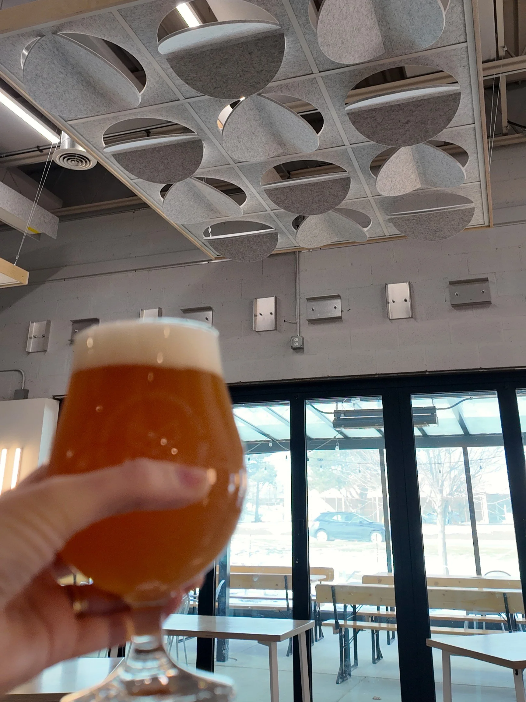 An hand holding a Mothfire beer in the foreground, in the back the taproom featuring the Leon x Askia Acoustic Island with an Oak frame and Heather Grey Luna tiles