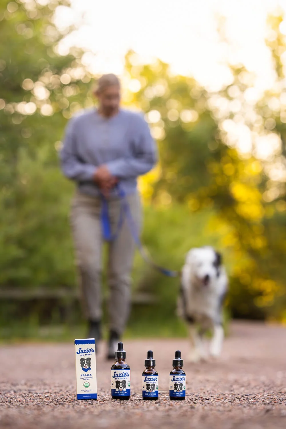 CBD products labeled 'Suzie's' in front of a person walking a dog on a trail during sunset.