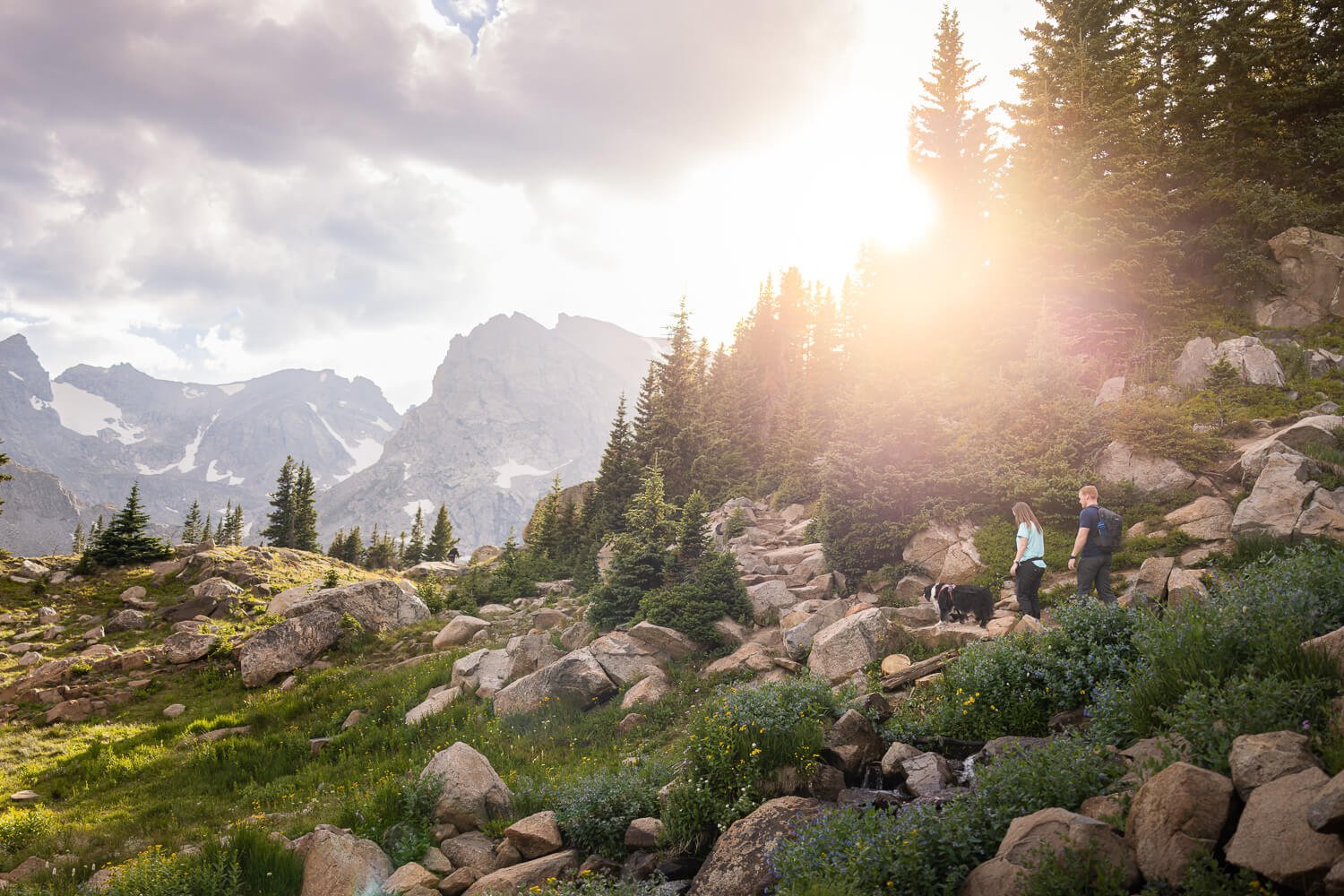 Lifestyle adventure pet photography capturing two people and a dog hiking on a rocky trail in the Colorado Rockies, with grand mountain peaks and a sun flare.