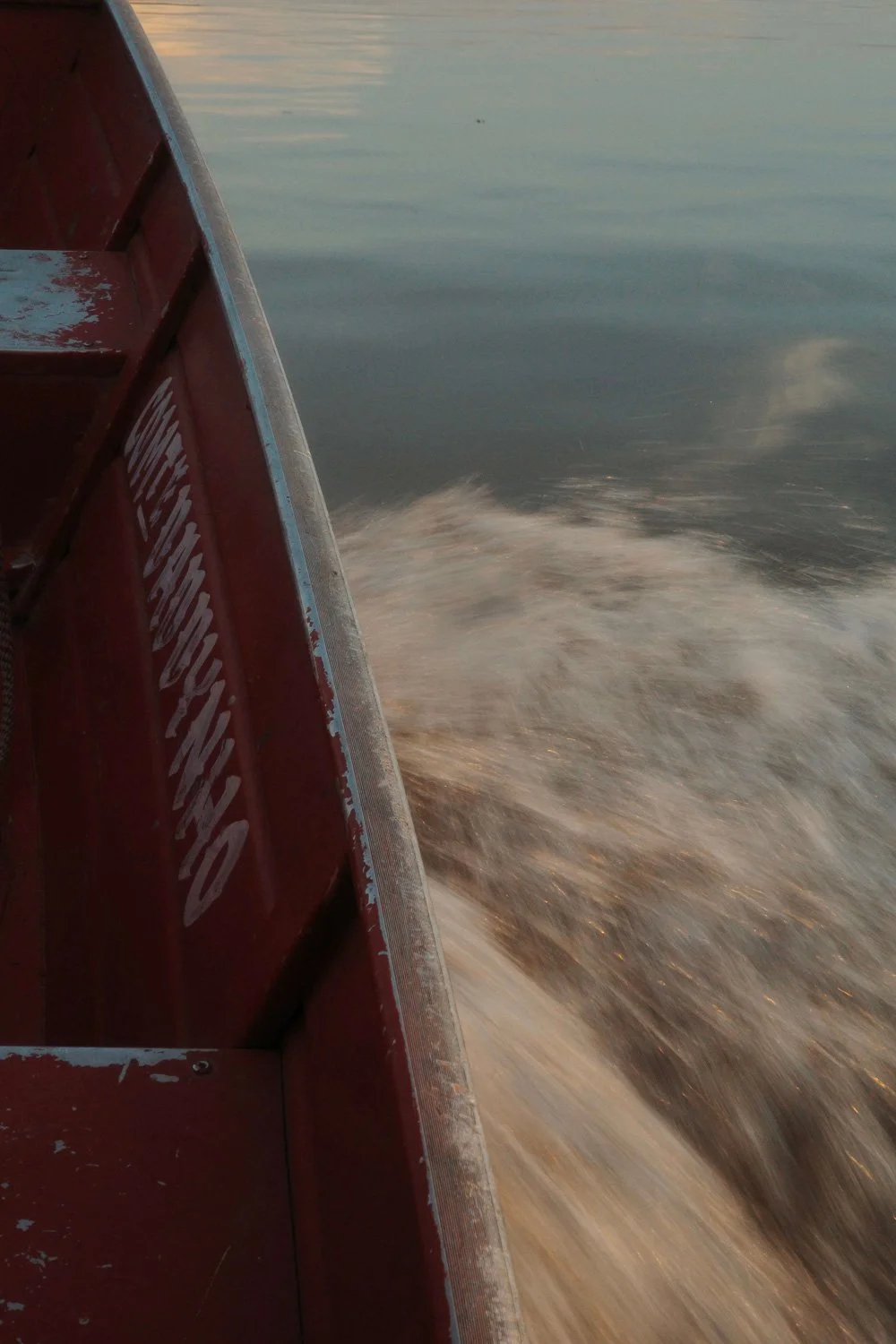 Red wooden boat hull cutting through the brown water of the Amazon River, with white spray churning in its wake.