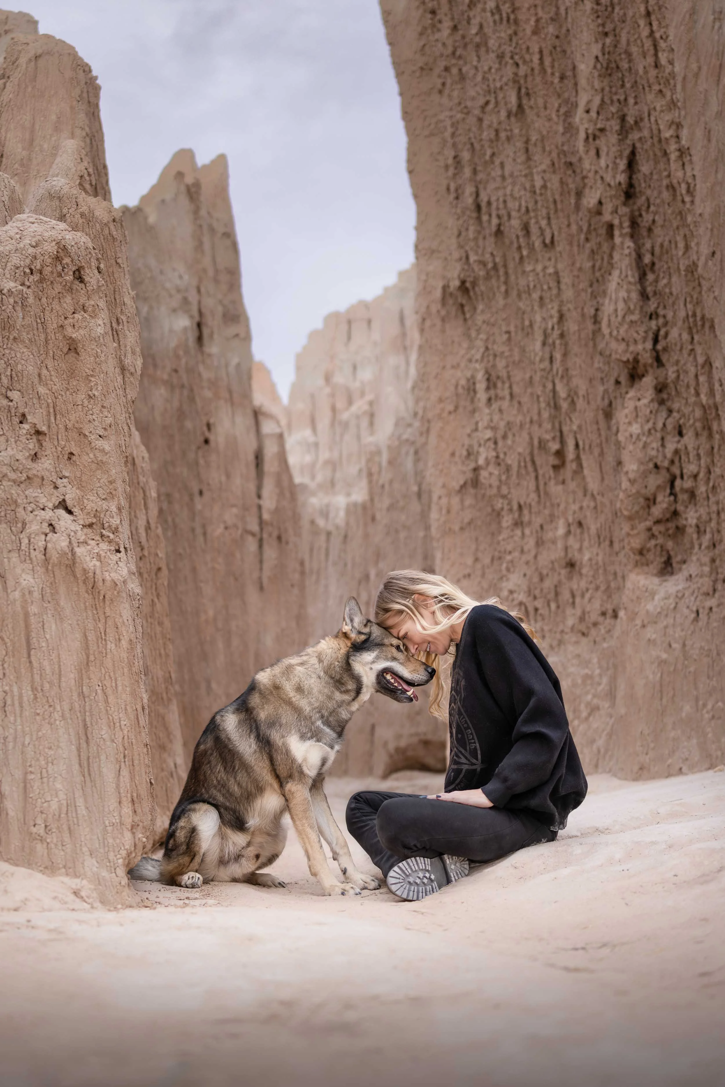 Woman sitting cross-legged on light sand sharing a sweet head-to-head moment with her dog inside a towering desert rock slot canyon.