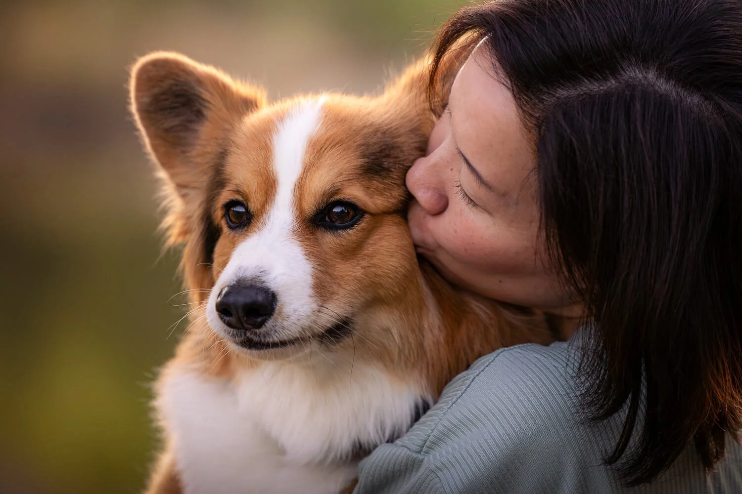 Close-up profile of a woman kissing her fluffy sable corgi on the cheek against a soft, warm backlight.